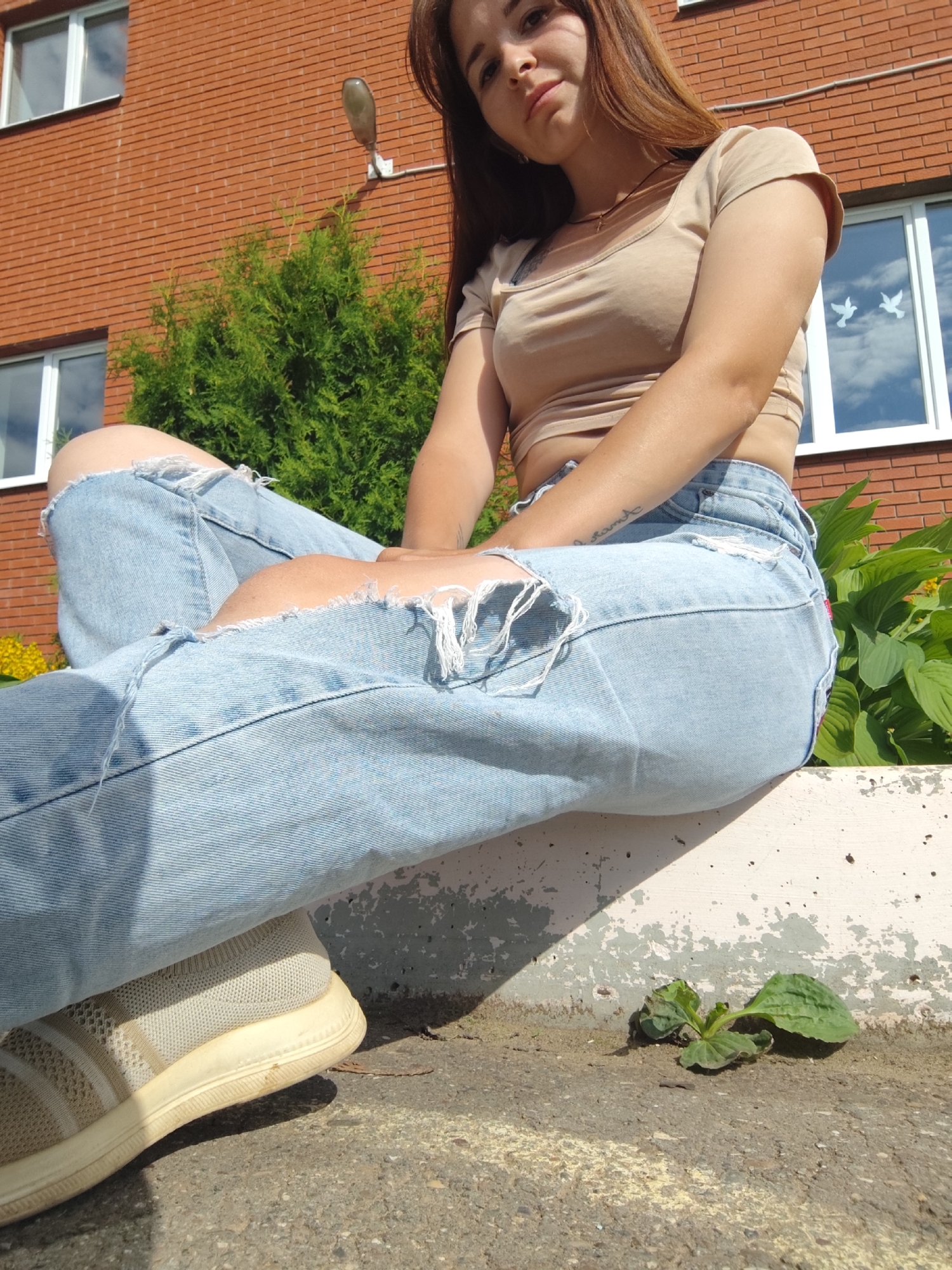 A young woman is sitting on a concrete ledge outdoors. She has long, brown hair and is wearing a light brown crop top and light blue ripped jeans. Her left leg is bent with her foot resting on the ground. She is wearing a light-colored sneaker on her left foot. Behind her, a brick building with two windows is visible, as is a green coniferous tree and green foliage. The scene appears to be in an urban or suburban setting, possibly in the city of Foky, Russia.