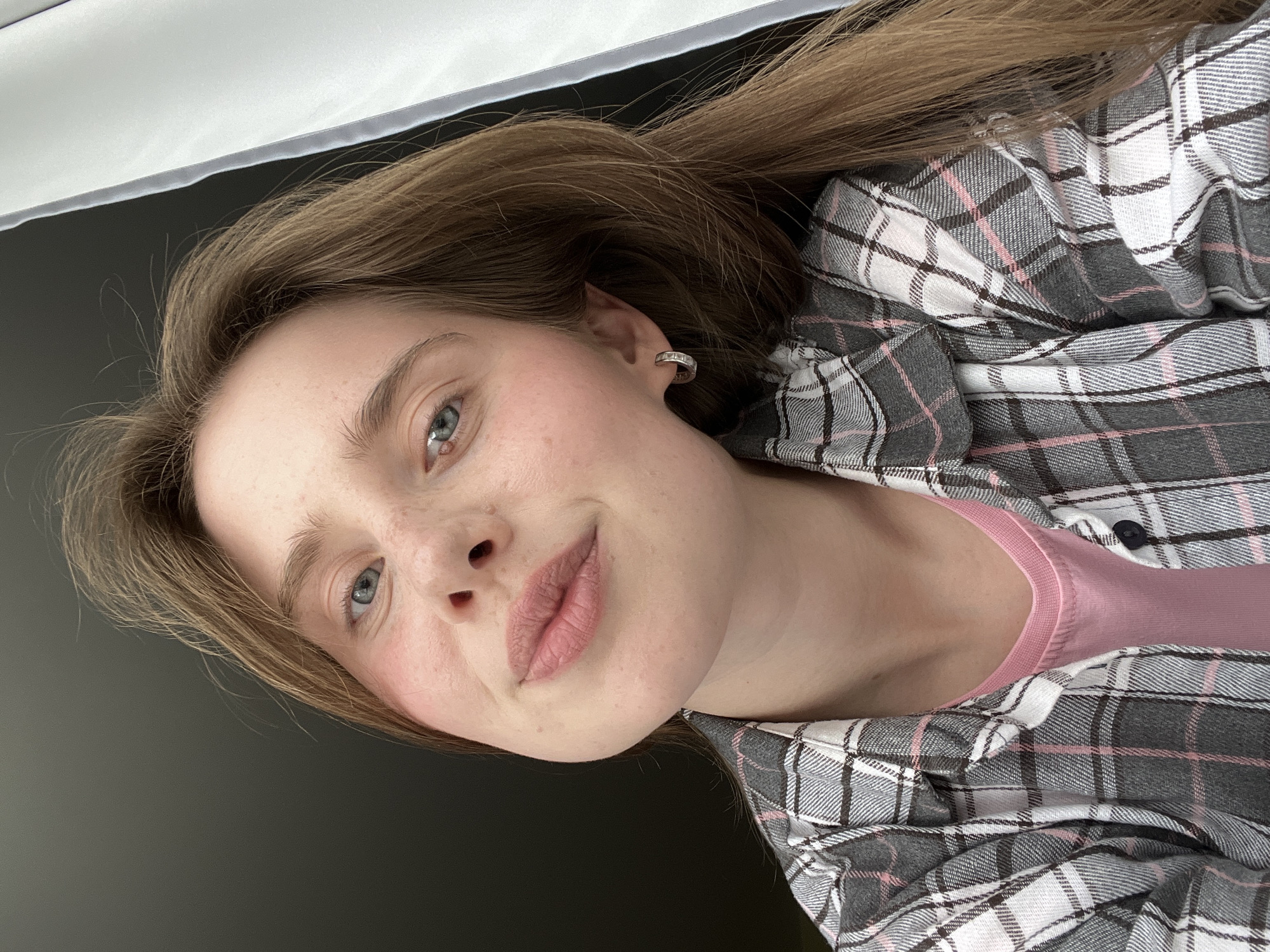 The image is a close-up selfie of a young woman with long, light brown hair. She is wearing a pink t-shirt under a grey and white plaid flannel shirt. Her skin is fair with some freckles and rosy cheeks. She has blue eyes and light pink lips, and is wearing a small silver hoop earring in her left ear. The background is dark on the left and a white curtain on the right. The image is taken indoors. There is no identifiable location in the city of Chaikovsky, Russia.