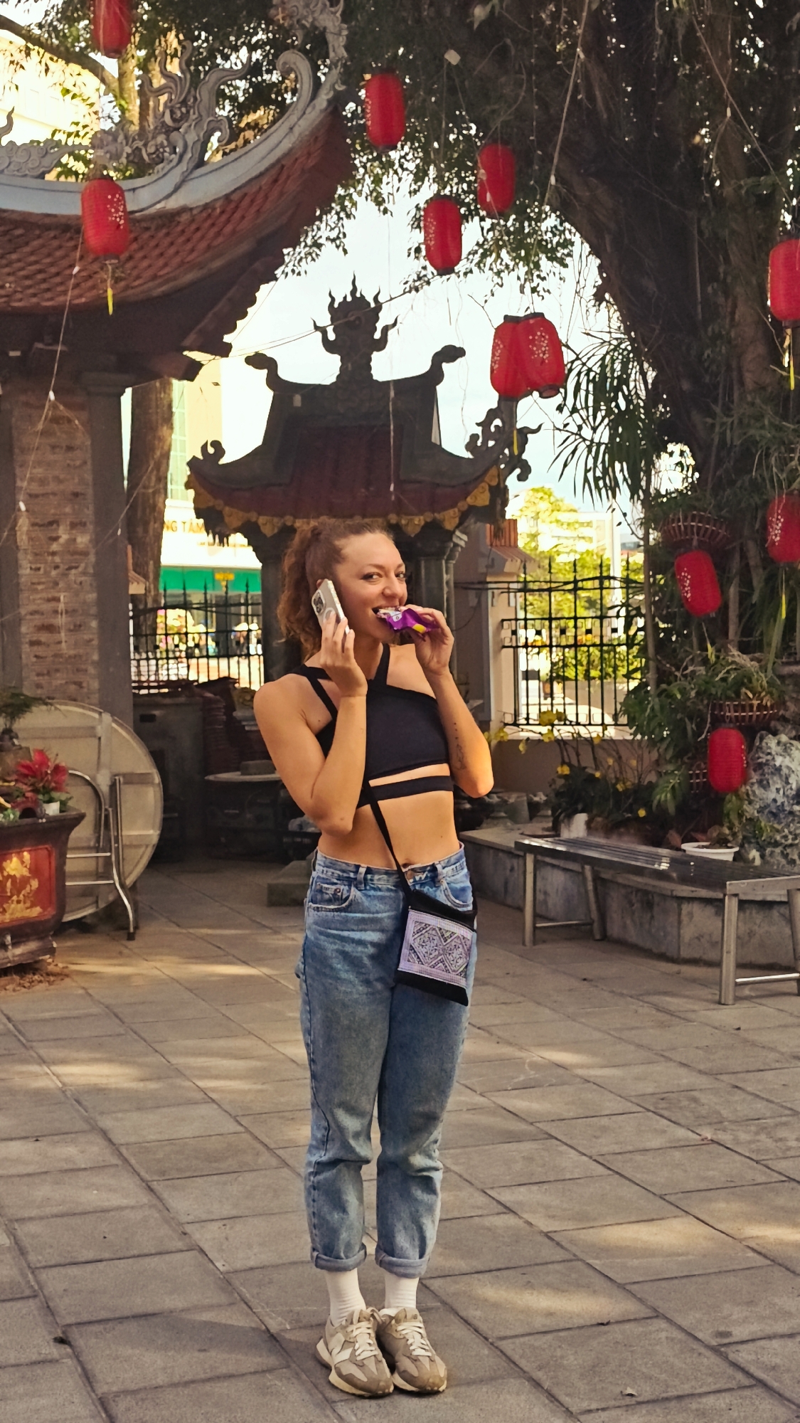 A full-shot captures a woman with red curly hair talking on her phone while eating, outdoors in Lao Cai Ward, Vietnam. The woman wears a black crop top, blue jeans, white socks, and tan sneakers. A purse hangs from her shoulder. Red lanterns decorate the scene, which includes traditional Vietnamese architectural elements like tiered roofs and a stone pathway. A fenced area with benches is visible in the background.