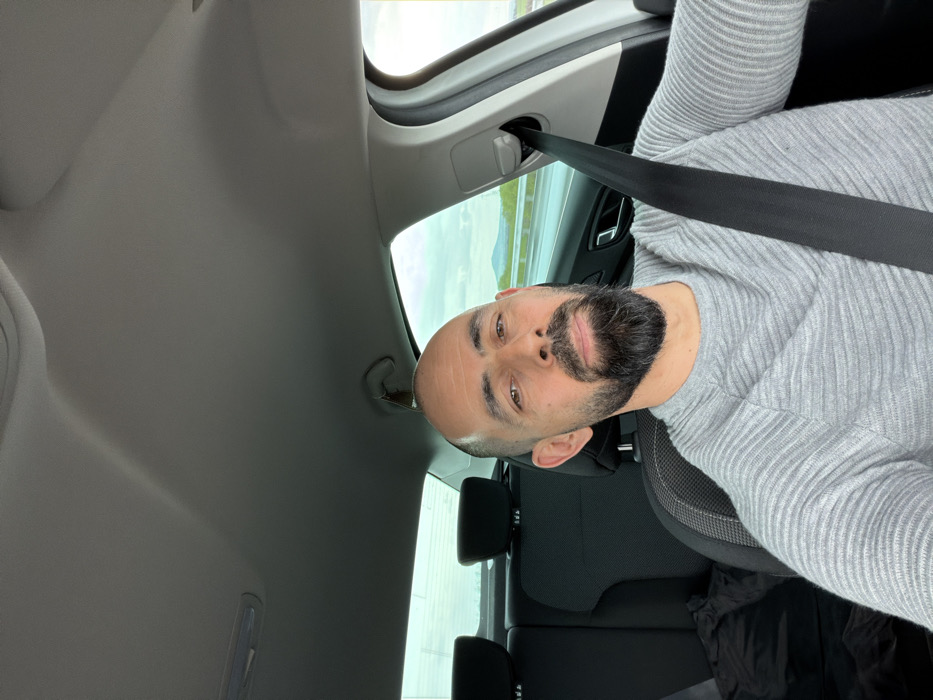 A bald male with a dark beard is depicted taking a selfie inside a vehicle. He wears a light grey, ribbed long-sleeved sweater and is secured by a black seatbelt positioned across his chest and lap. His gaze is directed towards the camera. The vehicle's interior features a light grey headliner. The rear passenger compartment is visible, showing two dark-colored headrests. A small object, possibly a hanger or air freshener, is suspended from the headliner near the driver's side rear door. Through the vehicle's windows, a highway is visible to the right and rear, suggesting movement. Distant hills or mountains under a partly cloudy sky with visible sunlight are present in the background. The scene is located within or in the immediate vicinity of Zetiovo, Bulgaria.