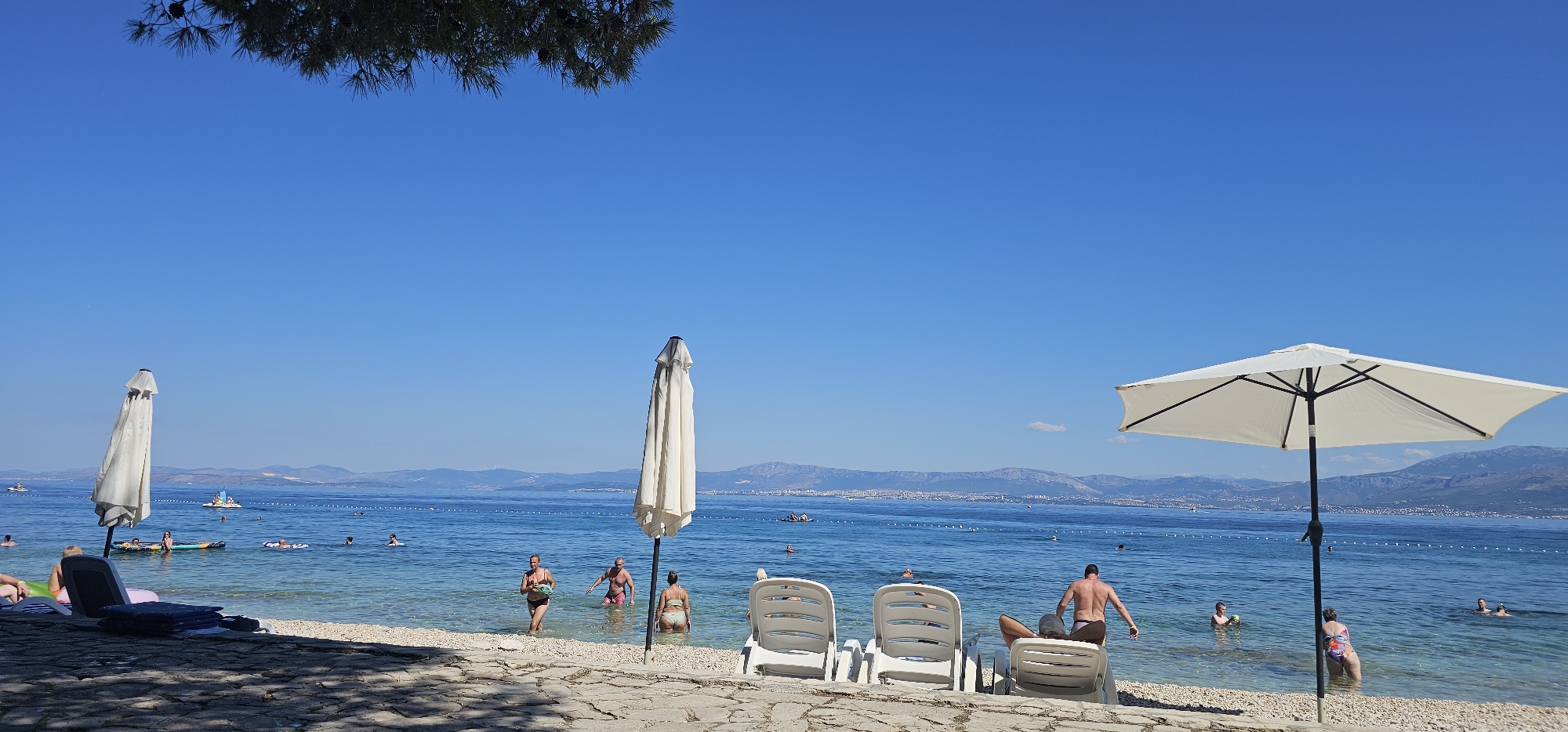 The image depicts a sunny day at a beach in Grad Supetar, Croatia. The foreground is a pebble beach with some stone paving, shaded by trees. Two white lounge chairs are positioned on the paved area, with one occupied by a person. A large white beach umbrella is open and casting shade on the area. In the mid-ground, the clear blue water of the Adriatic Sea stretches out, with several people swimming and floating on inflatable toys. Two closed white beach umbrellas are visible near the water's edge, and a white inflatable slide is in the distance. The background features a distant coastline with hills and mountains under a clear blue sky.