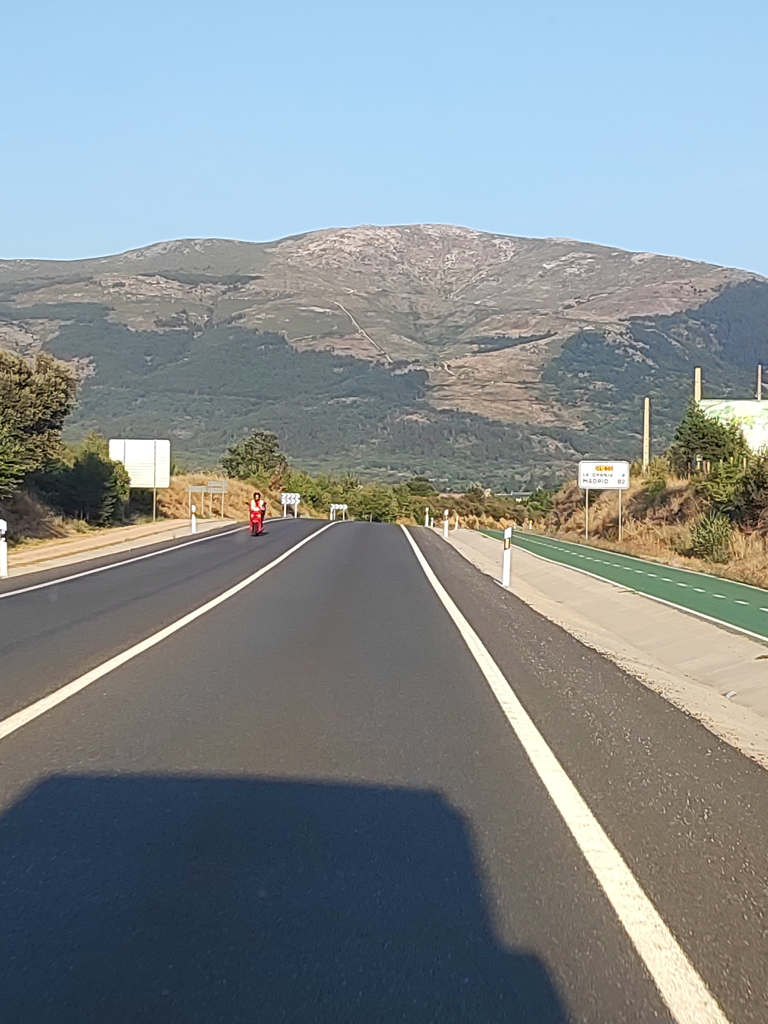 A person on a red scooter is traveling on a paved road with a white lane marker. The road is flanked by a green bike lane and a grassy shoulder with trees and vegetation. In the background, a large mountain range dominates the horizon, with a clear blue sky above. A white sign on the right side of the road indicates "LA GRANJA 4 MADRID 92," suggesting the location is near La Granja and Madrid. The image appears to be taken from a moving vehicle, as indicated by the shadow on the road in the foreground.