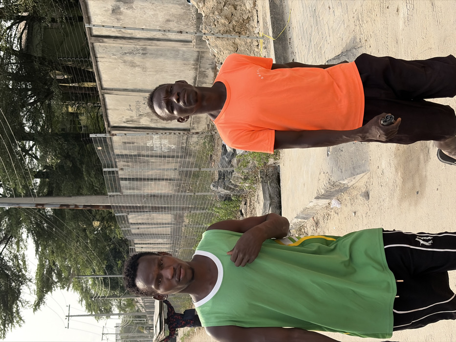 Two dark-skinned men stand side-by-side outdoors, facing the camera. The man on the right wears an orange shirt and dark pants, holding a small, dark object. The man on the left wears a green tank top with white trim and black pants with a white stripe. A wire fence and trees are visible in the background. The ground appears to be a mix of dirt and concrete. The image is rotated 90 degrees clockwise.