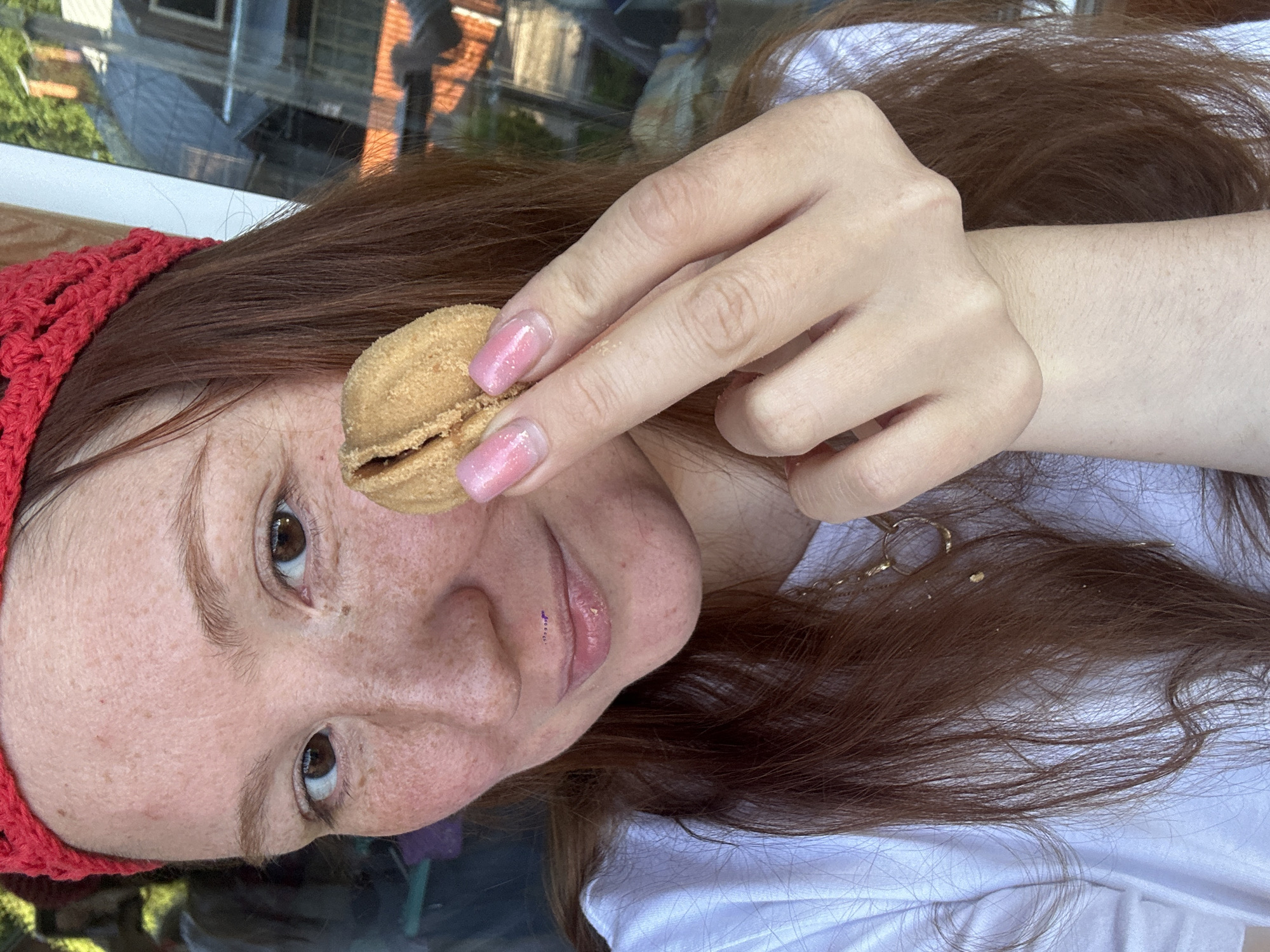 A woman with fair skin, freckles, and reddish-brown hair is holding a small, filled cookie to her face. She is wearing a red crocheted hat and a white t-shirt. The background shows a reflective window with trees and buildings. There is no definitive evidence in the image to pinpoint the location as Nizhny Novgorod, Russia.