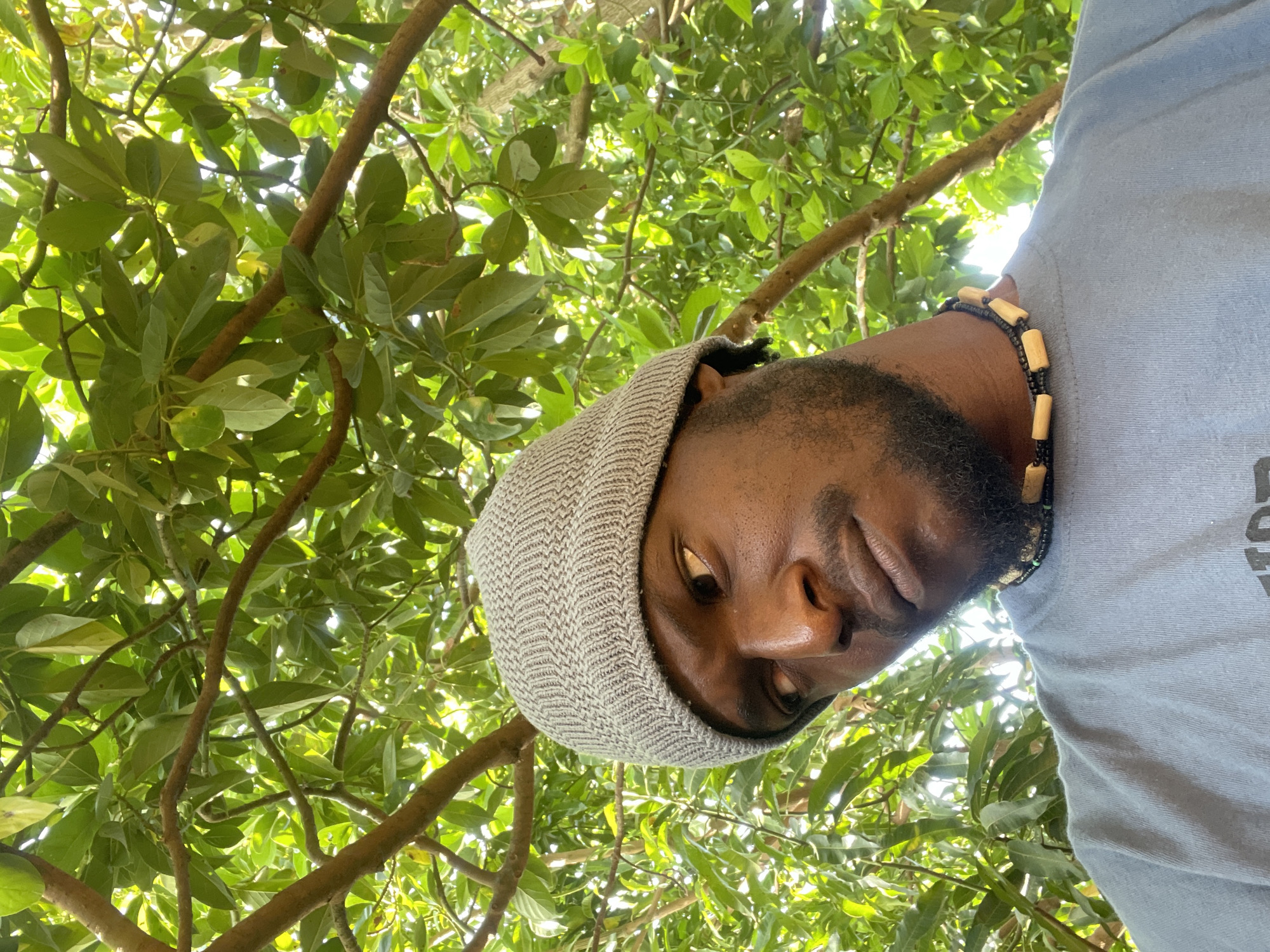 A man with dark skin, a full beard, and wearing a gray beanie and a necklace made of wood and black beads looks down at the camera. He is wearing a light blue shirt. Behind him are green leaves and tree branches, suggesting he is outdoors. The location is Jos, Nigeria.