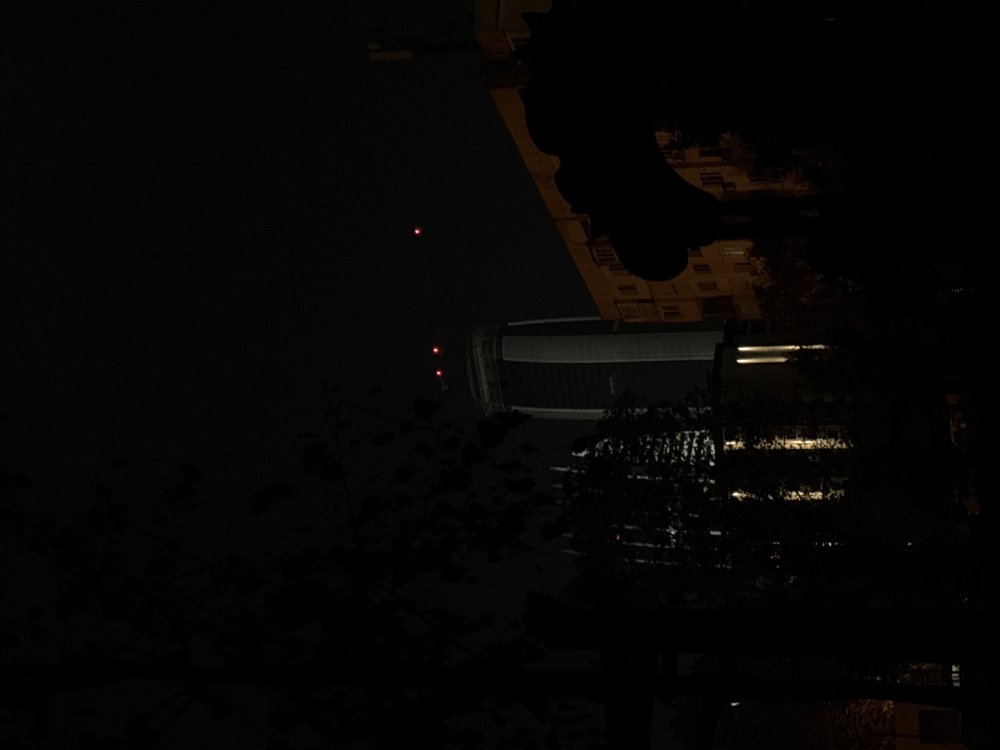 The image depicts a night scene in Sofia, Bulgaria. In the foreground, silhouetted trees obscure portions of buildings. A cylindrical structure with illuminated horizontal lines suggests modern architecture. A taller building is also visible, with lit windows arranged in a regular pattern. Near the top left, there are two vertically aligned red lights, possibly from a tower or antenna. The overall scene is dark, with the illumination primarily coming from the buildings and the distant red lights.