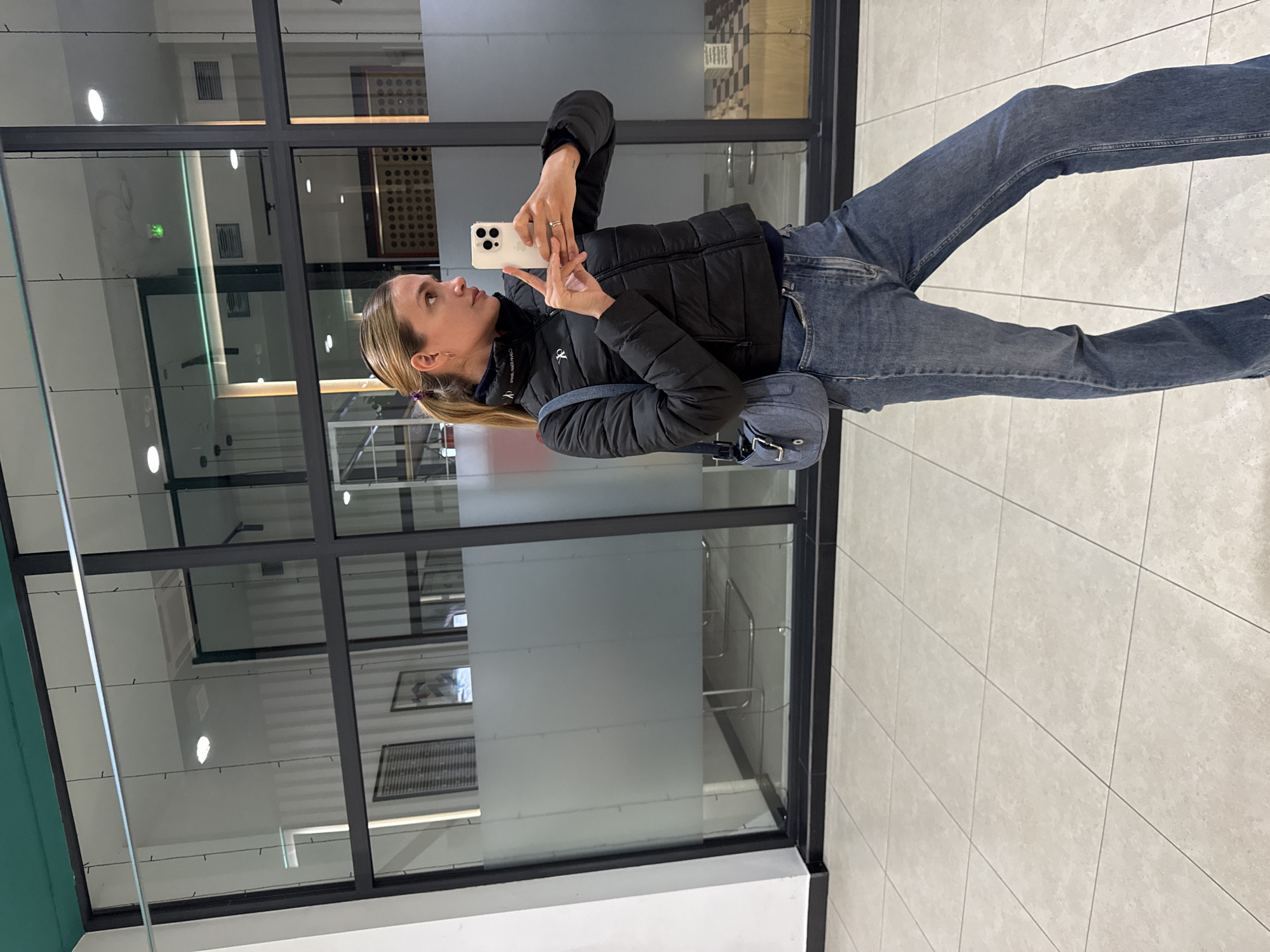 A woman in Tbilisi, Georgia, takes a selfie in front of a large, grid-patterned window. She is wearing jeans, a black puffer jacket, and a small denim purse slung across her body. She holds a white smartphone in her hand, pointing it at herself. The window behind her reflects the interior of the building and the outside environment. The floor is tiled.