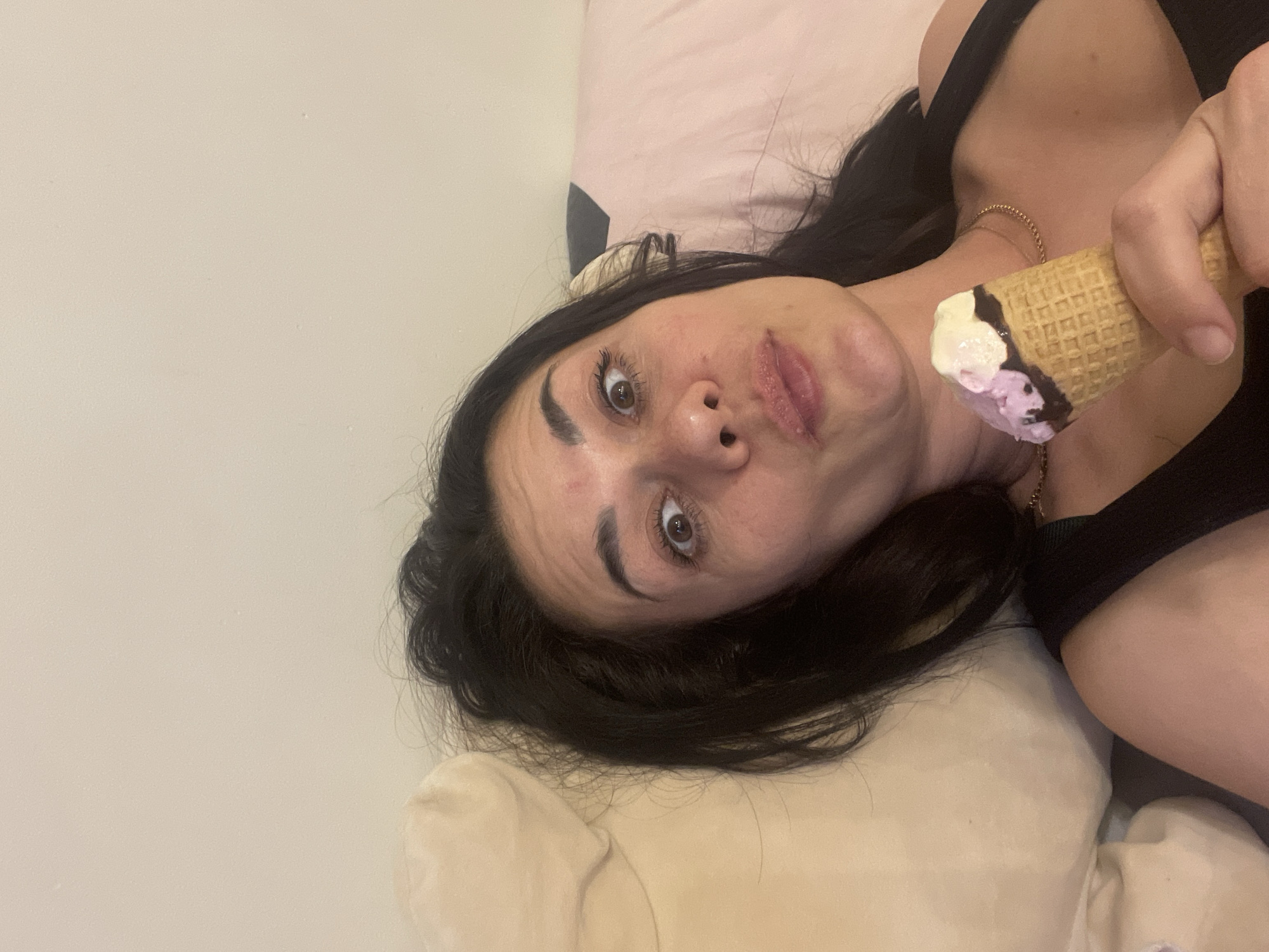 A woman is lying on a pillow, holding an ice cream cone. The ice cream is two-toned, with white and purple scoops and chocolate edging on the cone. She is wearing a black top and a gold necklace. She is looking at the camera with a slightly surprised expression. The background is a plain wall. The location is unknown.
