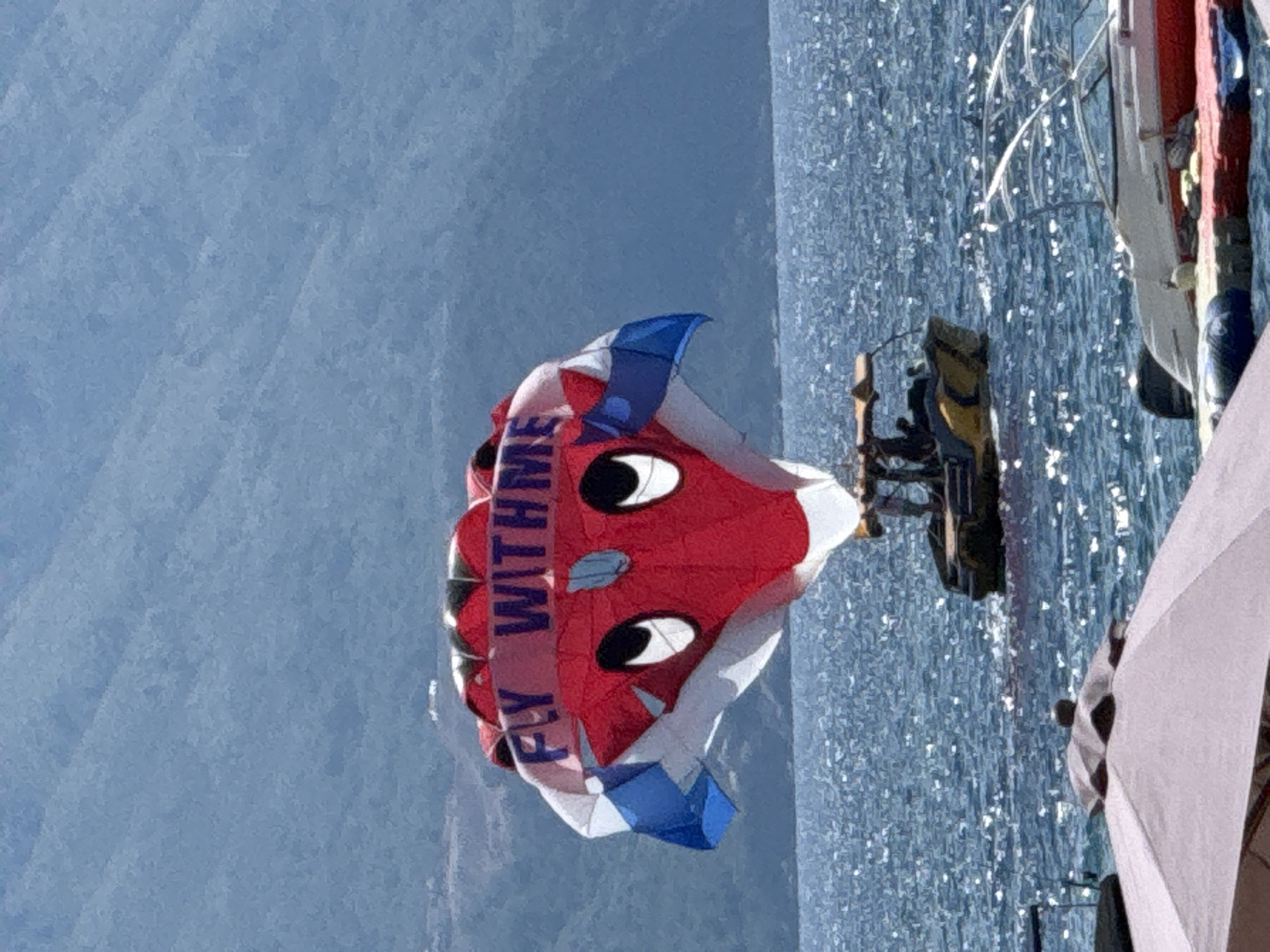 The image captures a parasail with a cartoon face and the text "FLY WITH ME" written in blue letters against a red background, suspended over a shimmering blue sea. The parasail is attached to a yellow boat with a canopy, on which two individuals are visible. In the background, a hazy, mountainous landscape dominates the scene, suggesting a coastal location. In the foreground, a portion of a light-colored umbrella and parts of two boats moored at a dock are visible. The scene appears to be set in Budva, Montenegro, a popular tourist destination known for its coastal beauty and water activities.