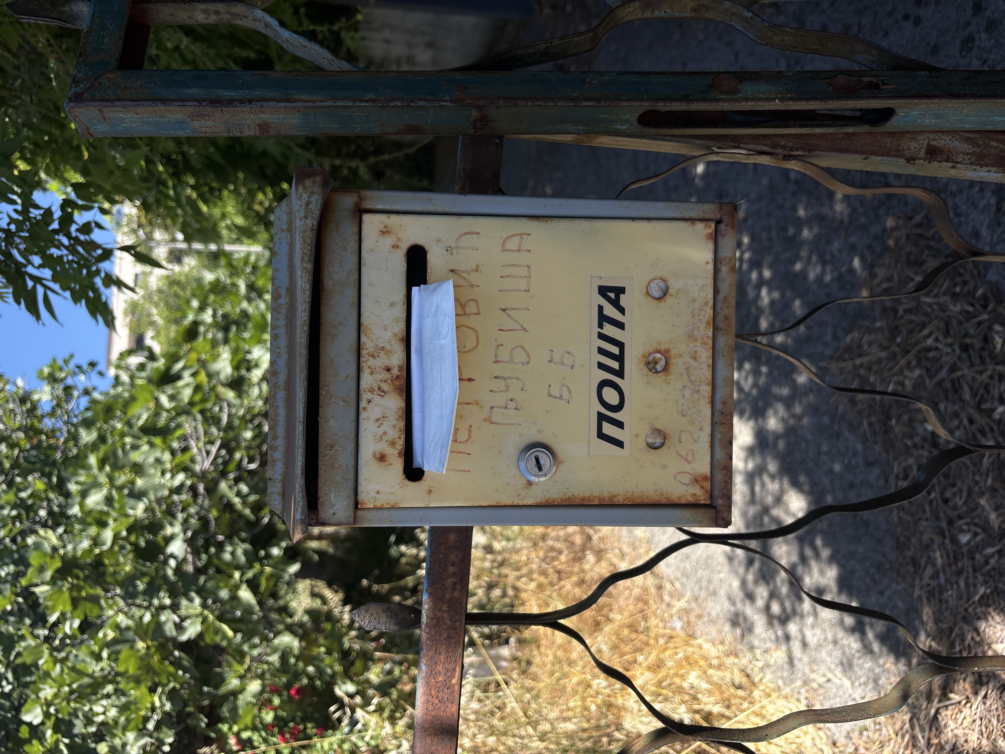An old, weathered yellow metal mailbox is mounted on a rusty metal gate in Boreti, Montenegro. The mailbox has a slot for mail at the top and a lock on the front. There is a white envelope partially inserted into the slot. The mailbox is adorned with handwritten text in Cyrillic script, the word "ПОШТА" (meaning "POST") in large black letters on a sticker, and what appears to be a phone number below. The gate is made of wrought iron and shows signs of rust and age. Behind the mailbox and gate, lush green foliage and trees are visible, along with a clear blue sky. The ground is covered with dry leaves and gravel. The overall scene suggests a rural or semi-rural setting, possibly a residential area, with an emphasis on the aged and functional nature of the mailbox.