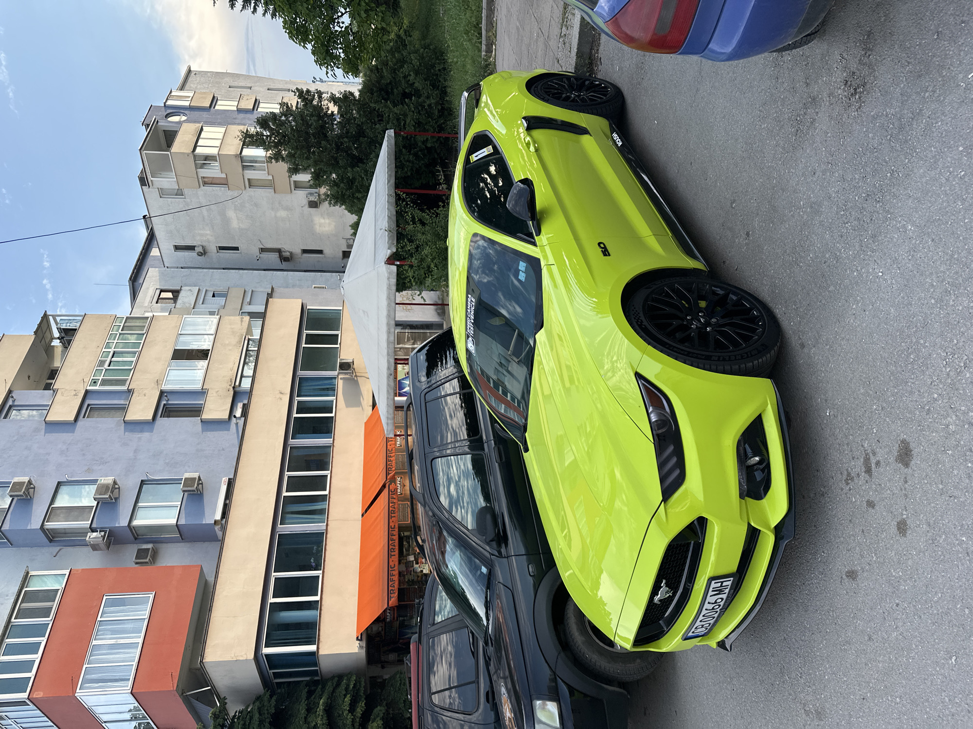 The image presents a daytime urban street scene in Veliko Tarnovo, Bulgaria, under a partly cloudy sky. The main subject is a striking neon green Ford Mustang, which appears to be a Roush variant, evidenced by "ROUSH" badging on its side fender. The car also features "5.0" badging, black multi-spoke wheels, and Michelin tires. Stickers on its rear side window show text including "TRAFFIC CAMERA" and "TEST VEHICLE". The front license plate reads "CB 0066 MH".
Behind the Mustang, a dark-colored SUV, likely a Nissan Xterra, is parked. The rear of a blue car is partially visible in the upper right corner of the frame. The background is dominated by several multi-story buildings with concrete facades, balconies, and numerous windows, some equipped with air conditioning units. An orange awning with the word "TRAFFIC" repeated is visible on one of the buildings, indicating a commercial establishment. Trees and green foliage are scattered throughout the scene. The overall impression is a static, contemporary urban environment with parked vehicles and no visible human activity.