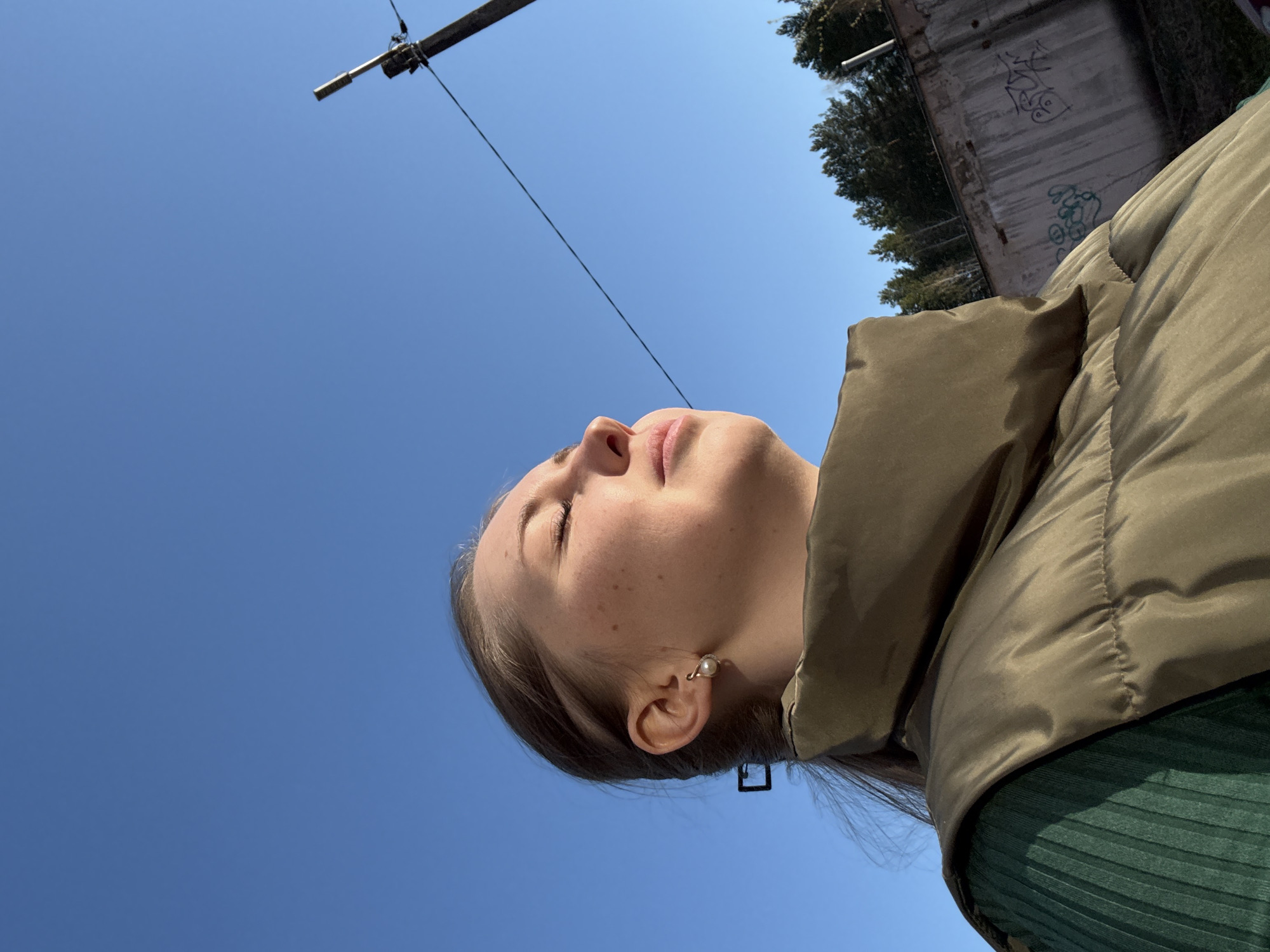A woman with fair skin and freckles is positioned in the lower-left quadrant of the image, with her head tilted upwards. She is wearing a pearl earring and a green ribbed top under a tan puffer jacket. Her eyes are closed, and she appears to be looking up at the sky.
The backdrop is a clear, bright blue sky, occupying the majority of the frame. In the upper-left corner, a utility pole is visible with a cable extending downwards. In the upper-right corner, a concrete building with graffiti and trees is also visible.
The location appears to be an urban environment, but the city is unknown.
