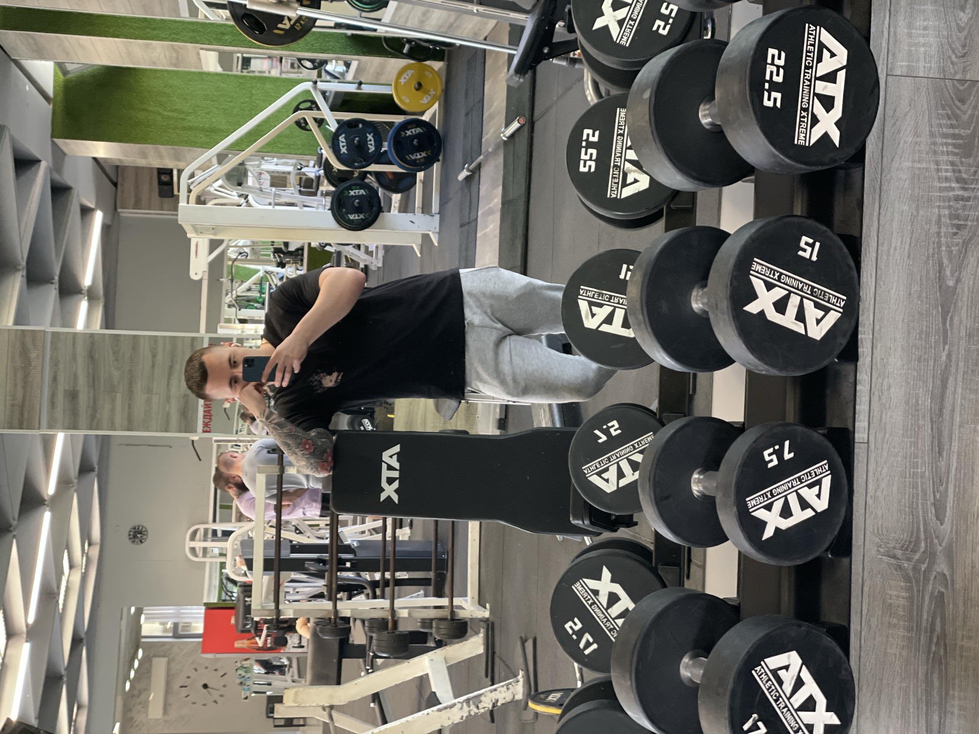 A man is taking a mirror selfie in a gym.
Location: Appears to be a gym in Asenovgrad, Bulgaria based on the presence of "XTX" branded equipment.
People:
- One man is taking a selfie.
- Other people are visible in the mirror reflection, engaging in workout activities.
Subjects:
- The man taking the selfie, clad in a black t-shirt and gray sweatpants.
Objects:
- Dumbbells of various weights (2.5, 7.5, 15, 22.5 kg) branded "XTX."
- A dumbbell rack.
- Weight lifting benches.
- Weight plates.
- A mirror reflecting the gym interior.
- Other gym equipment.
- A cellphone, held by the man.
Actions:
- The man is holding a cellphone and taking a selfie in front of a gym mirror.
- Other people are engaged in various gym activities in the background.
Interactions:
- The man is interacting with his cellphone to take a picture of himself in the gym setting.
Environment:
- The environment is a well-equipped gymnasium with weightlifting equipment. The interior includes a combination of gray flooring, reflective surfaces, and visible weightlifting apparatus.