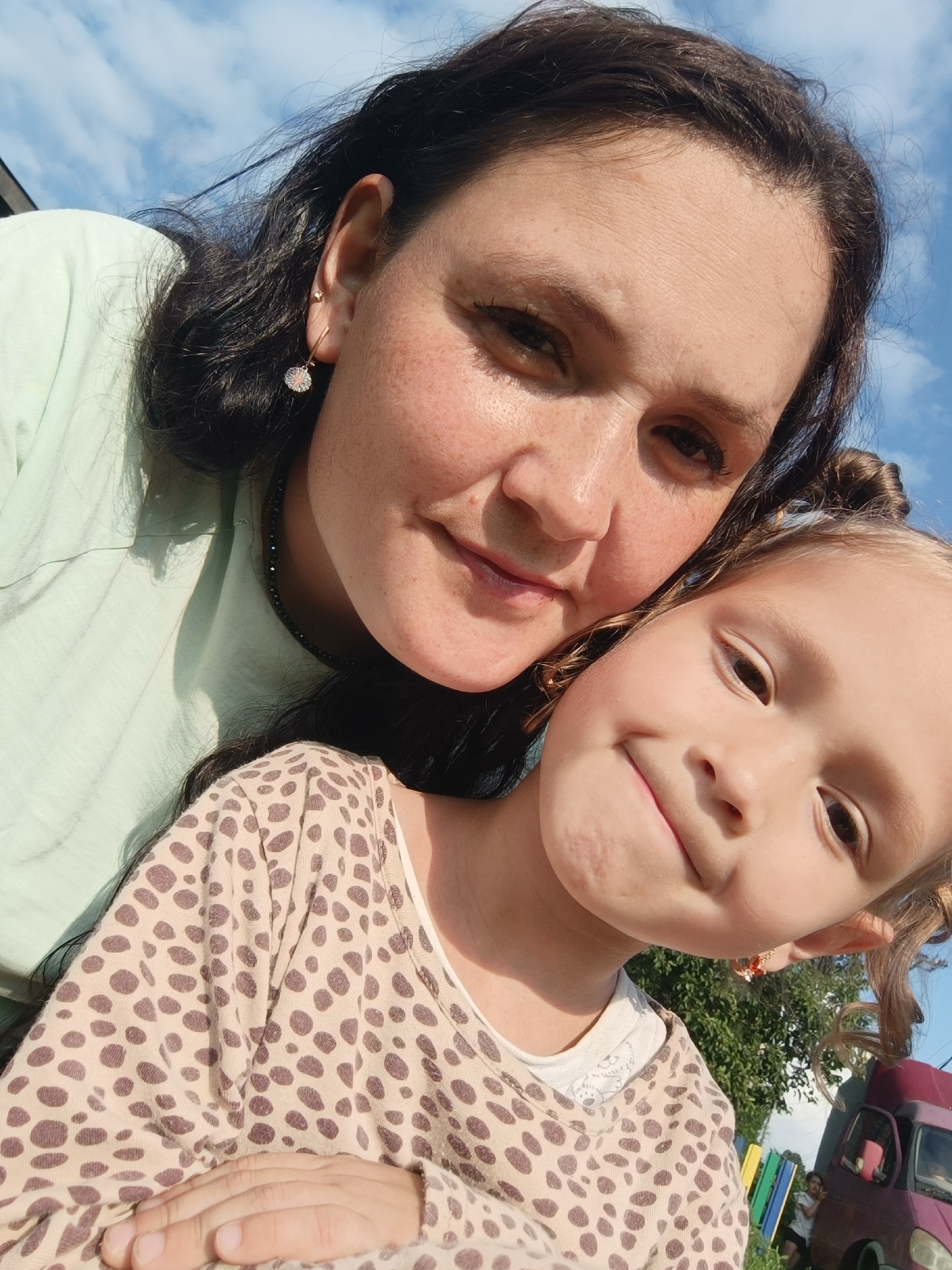 The image is a close-up, outdoor selfie of two females, likely mother and daughter, with their faces close together, smiling. The adult female, on the left, has dark hair and is wearing a light green shirt. She has freckles and is wearing a dangling earring. The younger female, on the right, has blonde hair tied up in two buns and is wearing a patterned shirt with small brown spots. She is also wearing an earring with red and gold accents. Their background consists of a bright blue sky with some scattered clouds, and foliage and a purple vehicle can be seen in the lower right corner, suggesting an outdoor, possibly rural or park-like setting. The image does not provide any information about the city of Karsha, Russia.