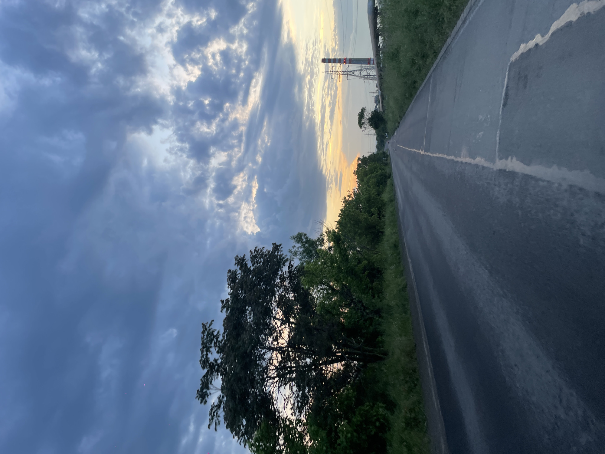 A slightly angled view of a paved road stretches into the distance, lined on both sides by green vegetation. Above the vegetation, a sky filled with gray and white clouds dominates the upper portion of the image. Near the horizon, a pale orange glow suggests either sunrise or sunset. Electrical towers are visible in the distance along the road. A large tree is featured in the lower-left corner. The road appears to be wet. No people are visible. The location cannot be determined to be the city of Burgas, Bulgaria.