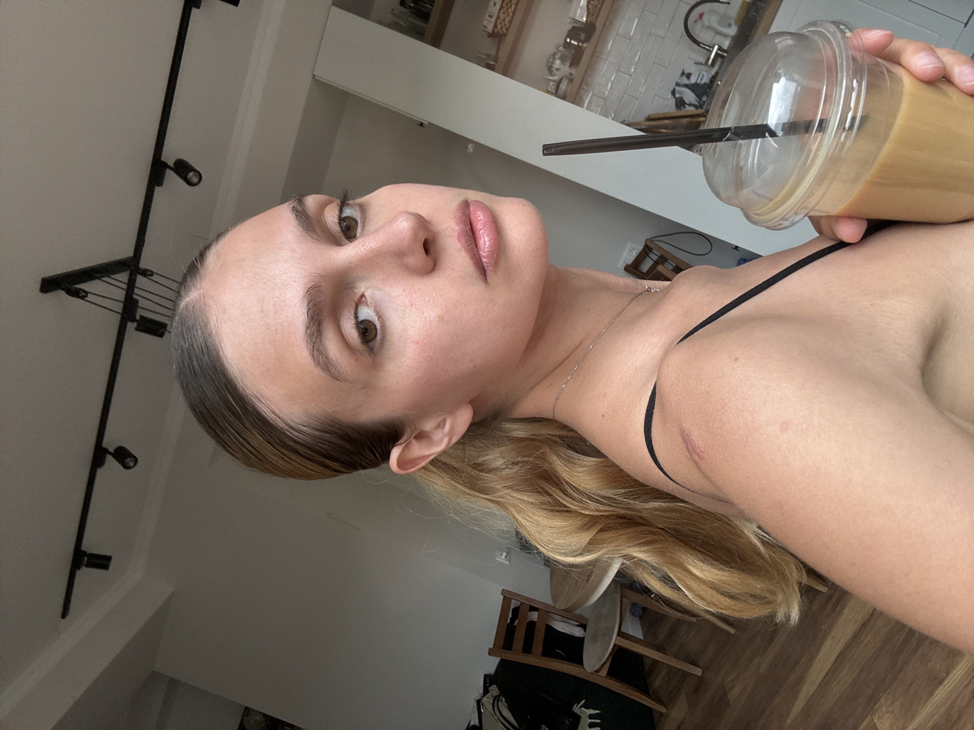 A woman is holding a plastic cup with a straw in an indoor setting. The cup appears to contain a coffee-based beverage. The woman has light brown hair and fair skin. She is wearing a thin black strap, likely from a garment. The background includes a track lighting fixture on the ceiling, a countertop area, and glimpses of furniture and flooring. The location is Unknown, Unknown.
