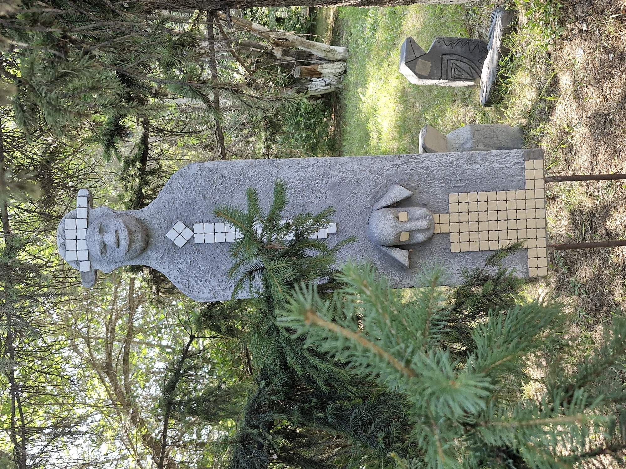 This is a photograph of an outdoor art installation located in Kokshetau, Kazakhstan. The primary subject is a large, grey, textured statue resembling a stylized human figure wearing a hat. The statue has a blocky, elongated torso decorated with white square mosaic tiles forming a geometric pattern. The head of the statue is simplified, with square eyes and a wide, smiling mouth. Below the head is a smaller, stylized dog-like face, also with square eyes, followed by a grid of small beige square tiles. The statue stands on two thin metal poles.
In the background, there are several other stone sculptures of varying sizes and abstract shapes. One is a dark grey, angular sculpture with carved geometric patterns. Another is a smaller, rounded grey sculpture. The sculptures are set amidst greenery, including grass, bushes, and mature trees with dense foliage, suggesting a park or garden setting. Sunlight filters through the tree branches, creating dappled light on the ground. The overall scene is one of an art display in a natural, outdoor environment.