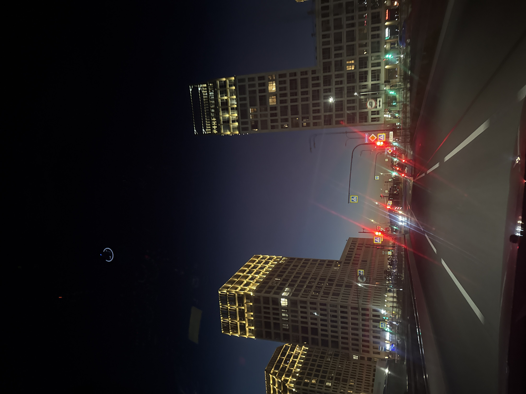 The image depicts a nighttime view from inside a vehicle driving on a multi-lane road in Saint Petersburg, Russia. The scene is dominated by modern, illuminated high-rise buildings on either side of the road. These buildings feature a contemporary architectural style with repetitive window patterns and are highlighted by warm yellow exterior lighting. Several traffic lights are visible, with some showing red and others green, indicating ongoing traffic flow. Pedestrian crossing signs are also present. The sky transitions from a deep blue near the horizon to black overhead, suggesting twilight or night. The road itself is paved and marked with white lane dividers. The overall impression is one of a developed urban environment at night.
