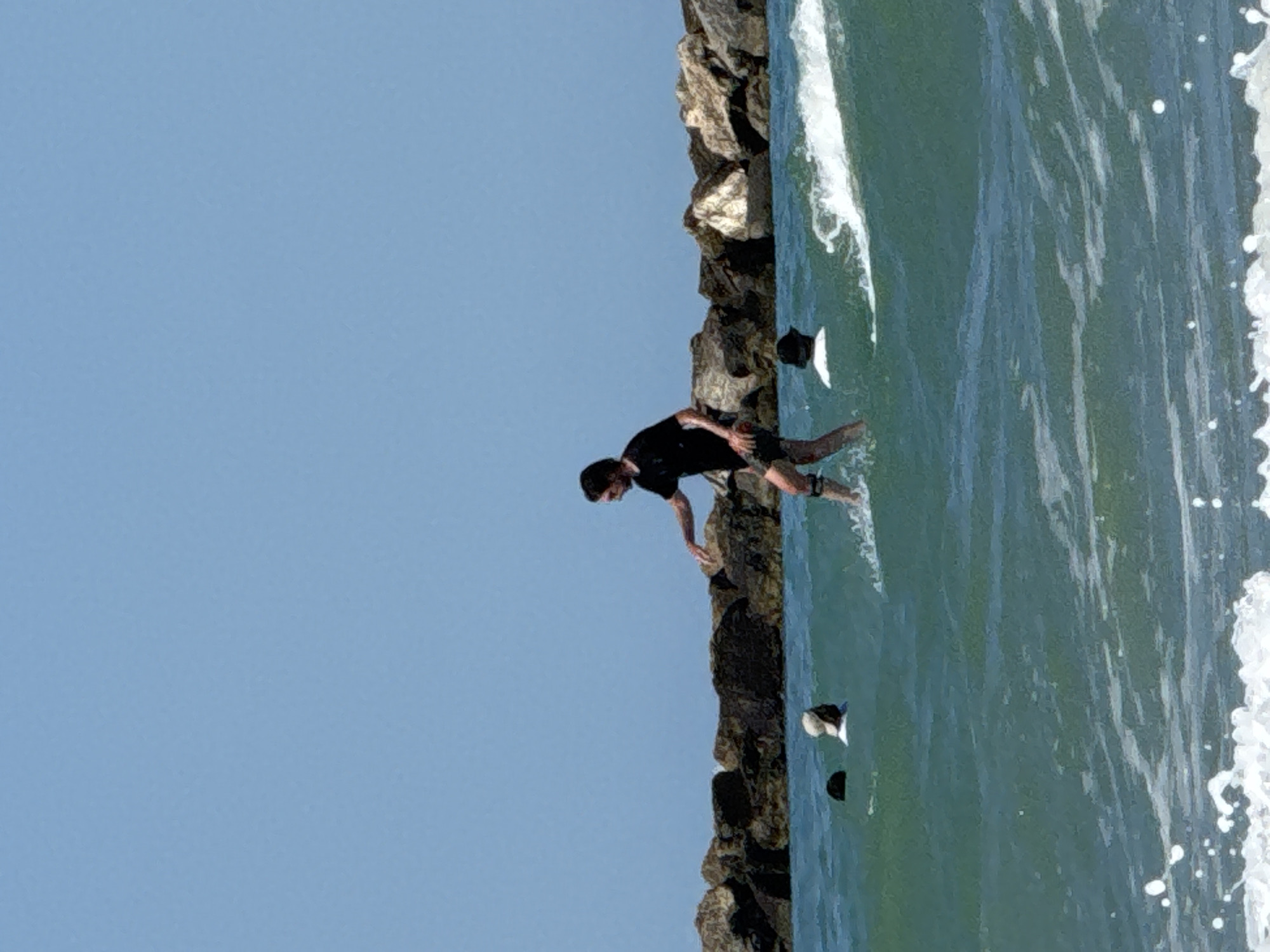 The image captures a sunny day at the beach in San Benedetto del Tronto, Italy, with a clear blue sky overhead. In the foreground, a person is shown surfing on a wave, wearing a black t-shirt and dark shorts. The surfer appears to be in motion, with water splashing around them. In the mid-ground, two individuals are partially submerged in the water; one is wearing a white cap and shirt, while the other is wearing a dark cap. In the background, a breakwater made of large rocks extends across the scene, separating the sea from what is likely a calmer area or the shore. The water is a vibrant turquoise, with small whitecaps and ripples indicating gentle wave activity. The overall atmosphere suggests a recreational day at the seaside.