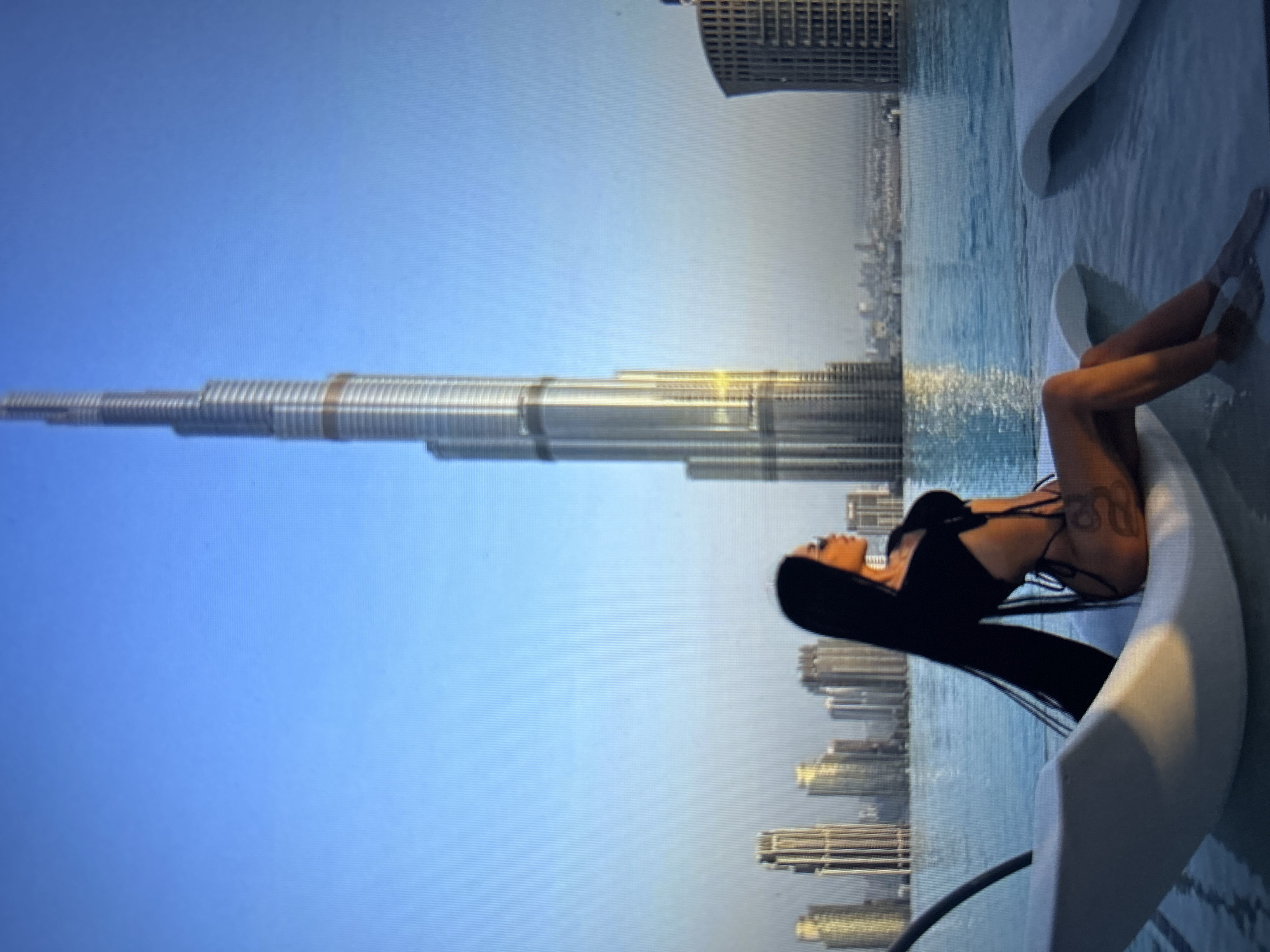 A full-shot, eye-level photograph captures a woman seated on a white lounge chair in an infinity pool. The woman has long, dark hair that flows down her back. She is wearing a black bikini and sunglasses. A tattoo is visible on her thigh. The pool water is blue, and the background features a cityscape with numerous tall buildings under a clear blue sky. The Burj Khalifa, a prominent skyscraper, is centrally located in the background. The scene is brightly lit, likely by natural daylight. The location is not Burgas, Bulgaria; it is Dubai, UAE, due to the presence of the Burj Khalifa.