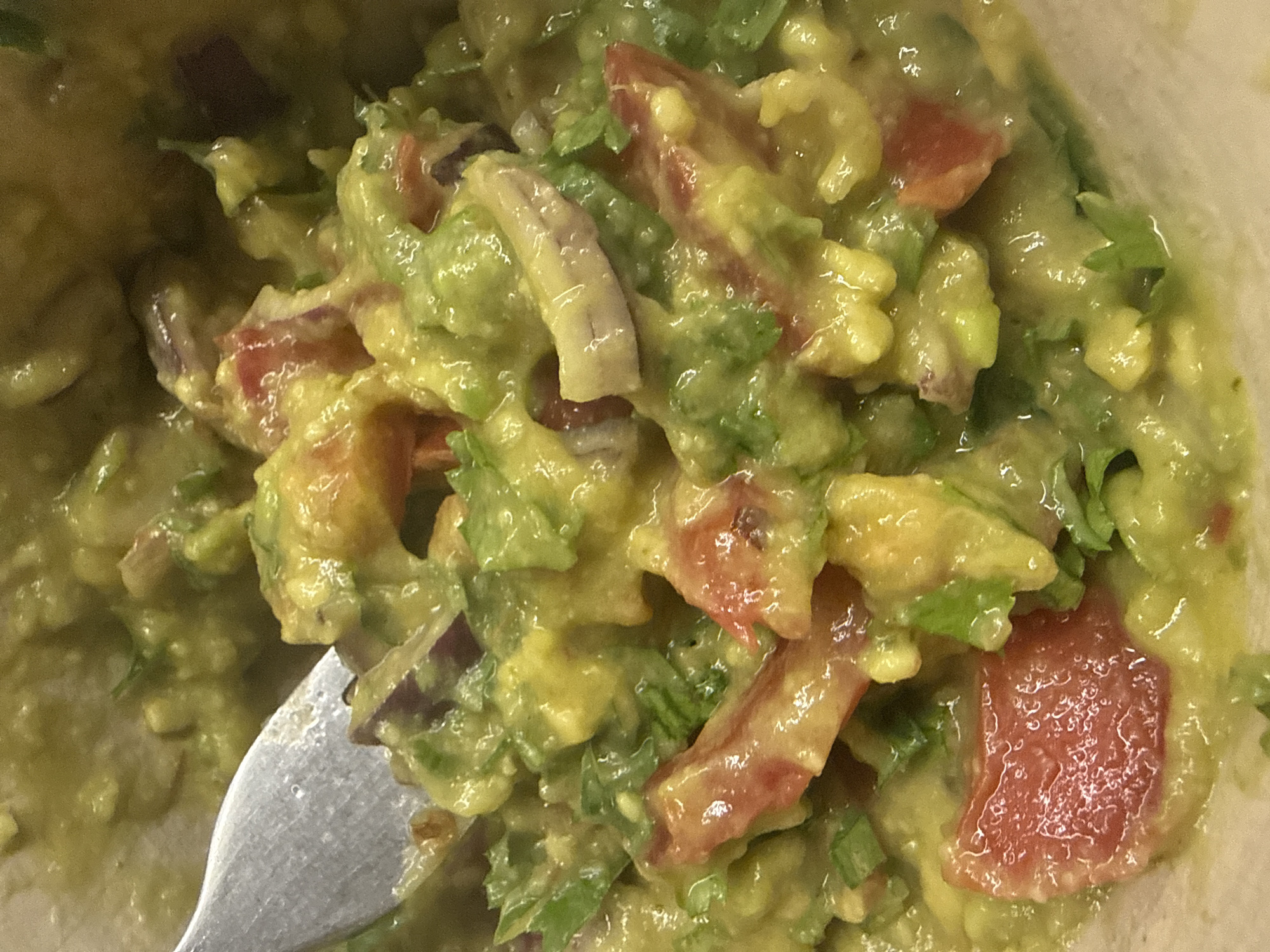 A close-up shot shows a fork piercing a bowl of freshly made guacamole. The guacamole is a vibrant green, with chunks of avocado, diced red tomatoes, and finely chopped red onion visible throughout. Sprinkles of green cilantro are also scattered within the mixture. The lighting is bright, highlighting the creamy texture of the guacamole and the fresh ingredients. The image is shot from a top-down perspective, with the fork positioned in the upper left quadrant of the frame. The background is blurred, drawing all attention to the food. The scene is located in a kitchen environment in the city of Sulitsa, Bulgaria.