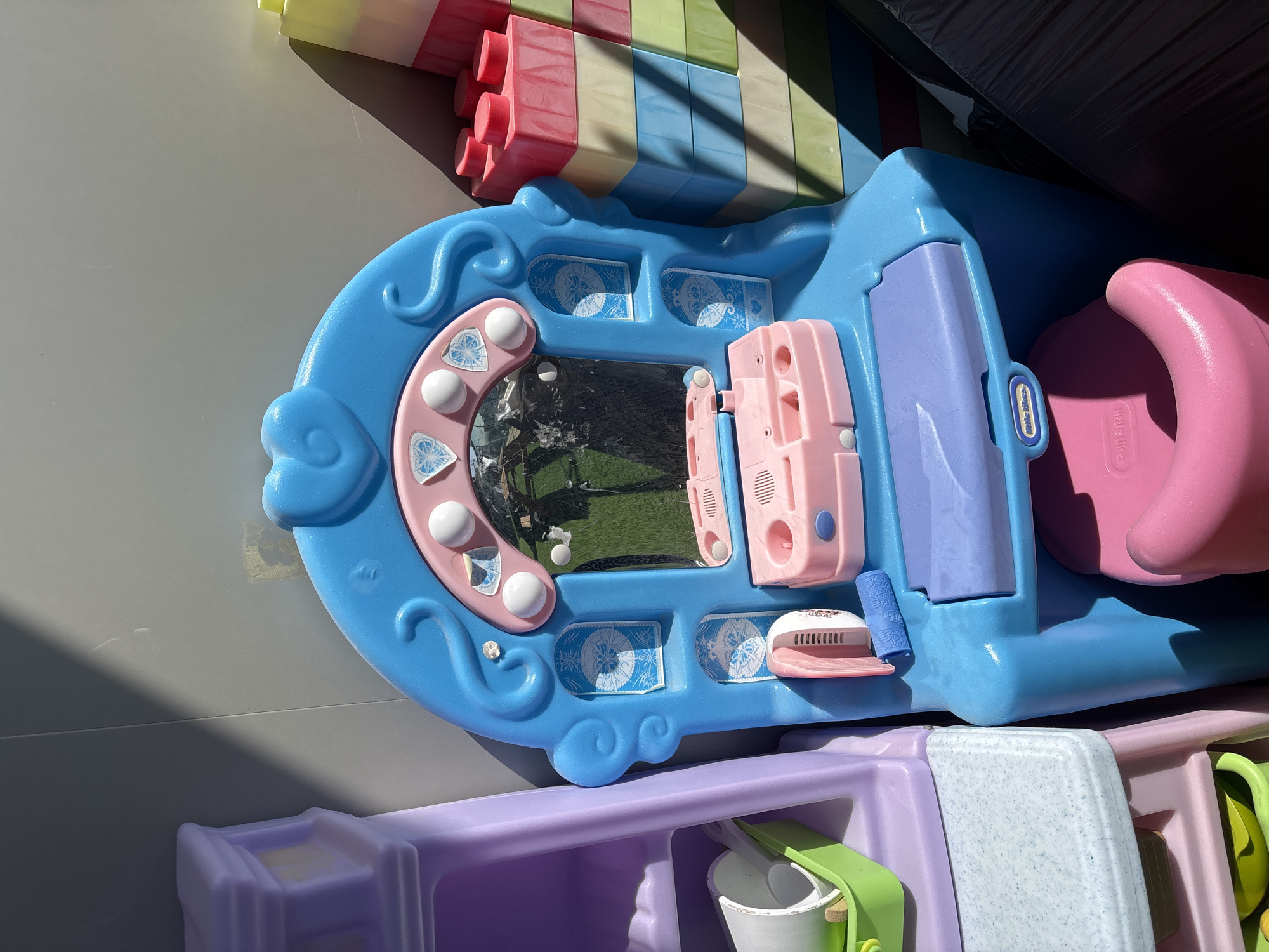 The image shows a child's play vanity and a stack of large, colorful building blocks, possibly from a toy set. The vanity is bright blue with pink accents, featuring a mirror, decorative elements, and what appears to be a play makeup station. To the right of the vanity is a tower of oversized building blocks in various pastel colors, including pink, yellow, blue, and green. In the background, a light gray wall is visible, with a distinct shadow cast by sunlight, suggesting an outdoor setting. The overall scene suggests a play area or storage area for children's toys, possibly in a residential or recreational setting within Dubai, United Arab Emirates. There are no people or animals present in the image.