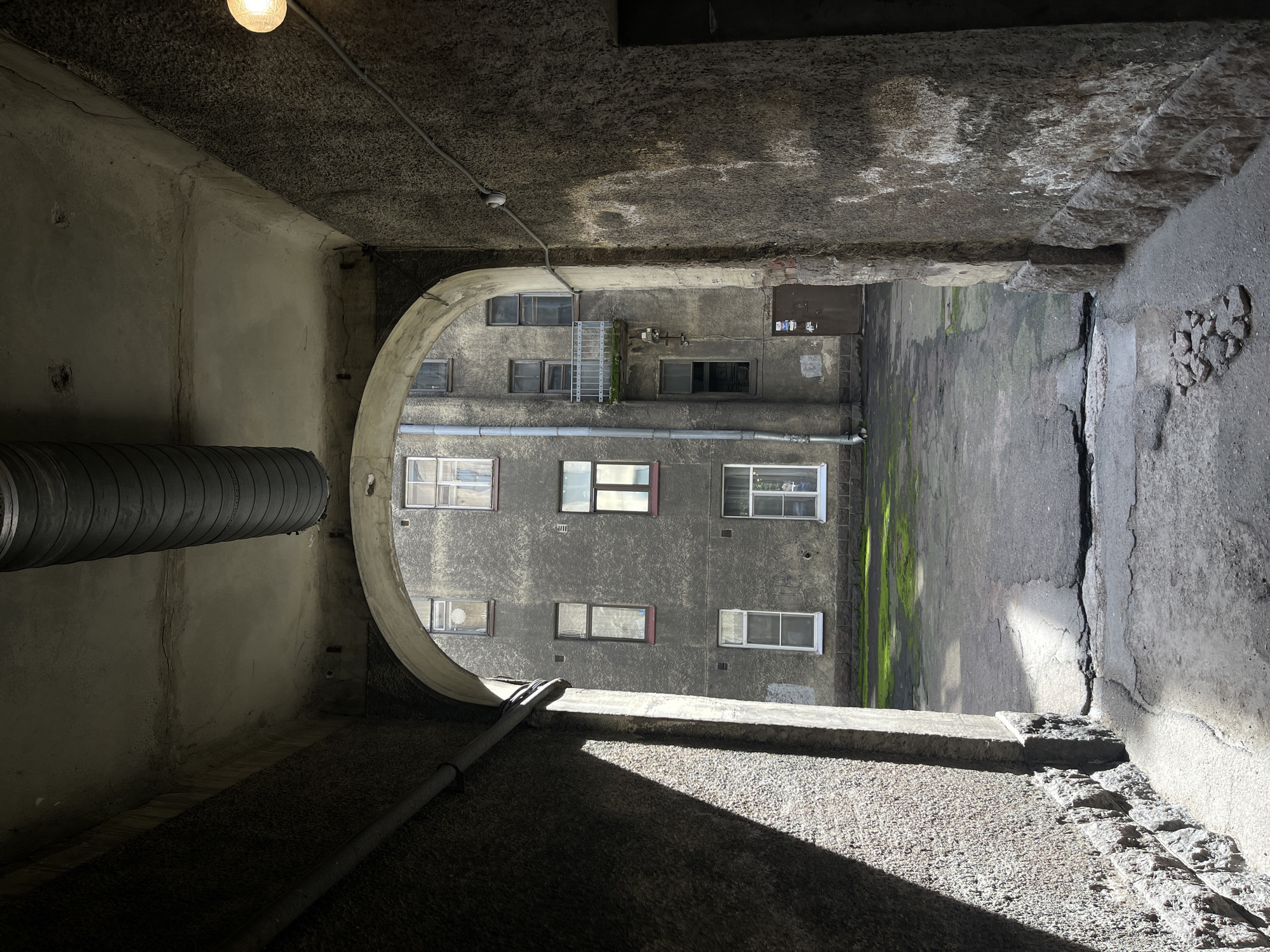 The image depicts a view through an arched entryway into a courtyard. The entryway is dark and textured, with a large corrugated pipe running along the ceiling. On the right side of the entryway, a lightbulb is visible. The courtyard is paved and shows signs of wear and tear, with cracks and patches. Green moss grows in patches on the ground. The buildings surrounding the courtyard are multi-story and appear aged, with numerous windows. A small balcony with a metal railing is visible on one of the buildings. A brown door is present at ground level on one of the buildings. The overall scene suggests an older urban environment. The architecture and building materials are consistent with those found in Vyborg, Russia, which has a rich history influenced by Swedish and Finnish periods.