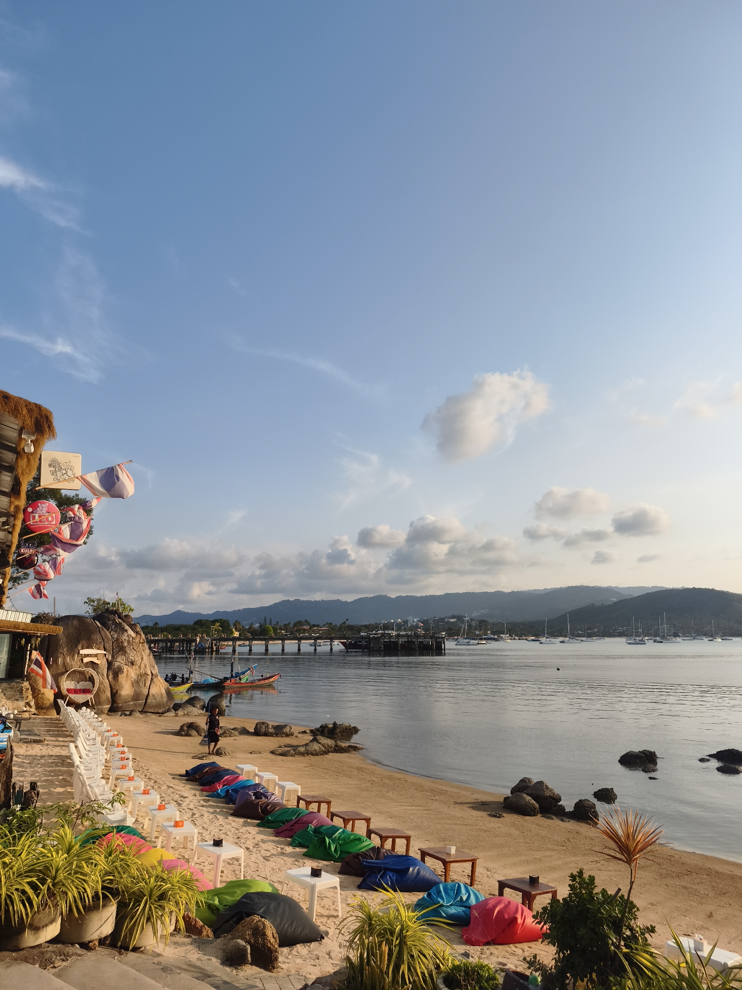 This photograph shows a coastal scene in Baan Plai Laem, Thailand. The sky, which covers the upper portion of the frame, is clear blue with sparse white clouds. Below, the sea is calm and reflects the sky. In the distance, mountains are visible.
The foreground features a sandy beach with rows of colorful beanbag chairs and small tables, presumably for patrons. To the left, a structure with a thatched roof and various signs and decorations stands, possibly a beachside bar or restaurant. A row of white chairs is lined up near the edge of this structure. Potted plants are at the bottom left. A pier is visible in the background, with several boats and sailboats floating in the water. A person is standing on the beach near the water.