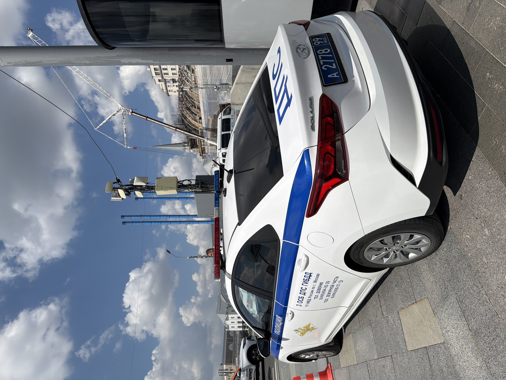 The image shows a white police car, a Hyundai Solaris, with blue and white markings and a flashing blue and red light bar on the roof. The car is parked on a paved sidewalk in Moscow, Russia. Text on the car indicates it belongs to the "ПОЛИЦИЯ" (Police) and "ДПС" (Road Patrol Service). A license plate reading "A 2778 99 RUS" is visible. In the background, construction cranes and buildings are visible under a blue sky with scattered clouds. Another white vehicle with "Яндекс Драйв" (Yandex Drive) branding is parked behind the police car. The scene appears to be a public urban environment.