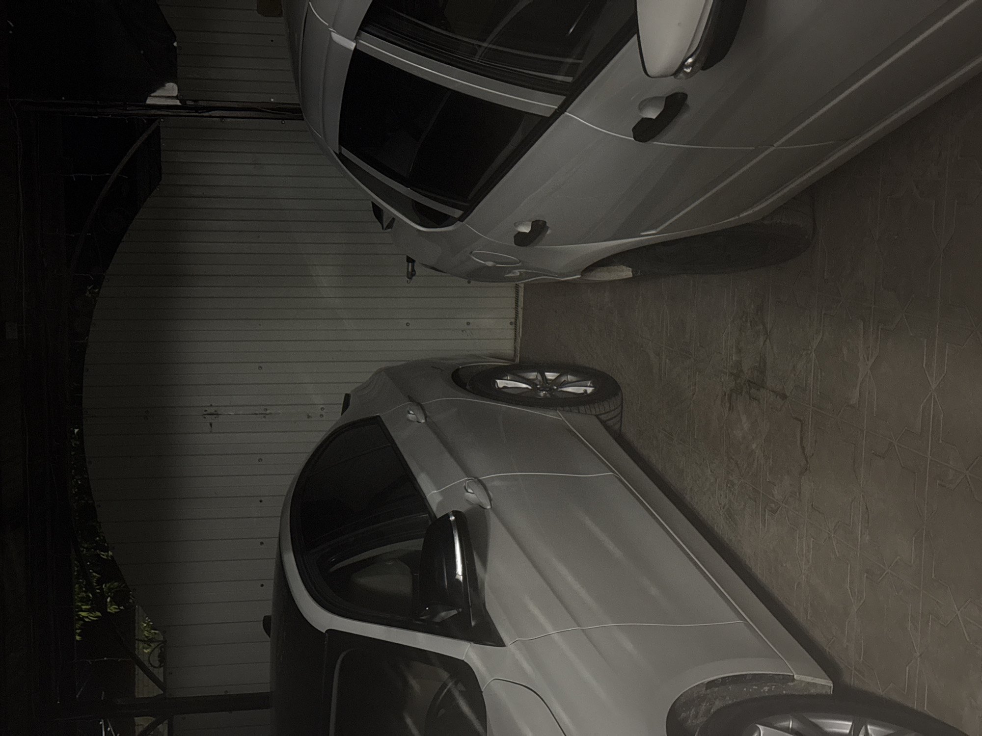 Two light-colored sedans are parked side-by-side in a covered area with a corrugated metal wall behind them. The car on the left is mostly visible from the front quarter, showing its driver's side door, side mirror, front wheel, and a portion of the roof. The car on the right is visible from its rear quarter, showing its passenger side rear door, rear wheel, and trunk. The ground is paved with interlocking concrete tiles. The scene appears to be a residential garage or a parking area in Mikhaylovsk, Russia, possibly at night due to the low lighting and dark shadows. No people are present in the image.