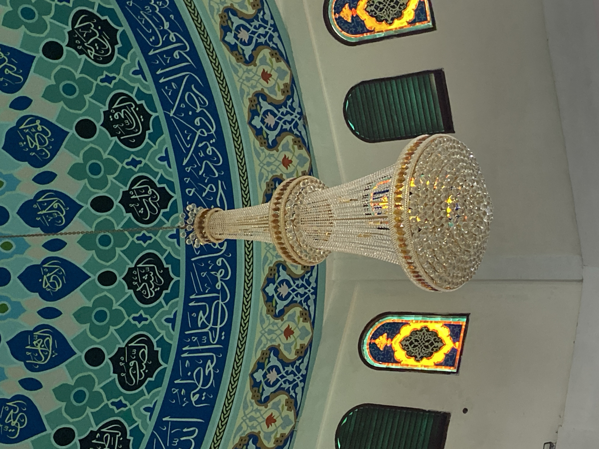 The image shows the interior of the Baiturrahman Grand Mosque in Lhokseumawe, Indonesia, specifically focusing on the ornate ceiling and a chandelier. The ceiling is decorated with intricate Islamic calligraphy and geometric patterns in shades of blue and teal. Below the ceiling, a large, multi-tiered crystal chandelier hangs, sparkling with light. Three arched windows are visible along the wall, two of which have stained glass with yellow and blue designs. One window on the left appears to have green louvers. The overall impression is one of grandeur and religious artistry.