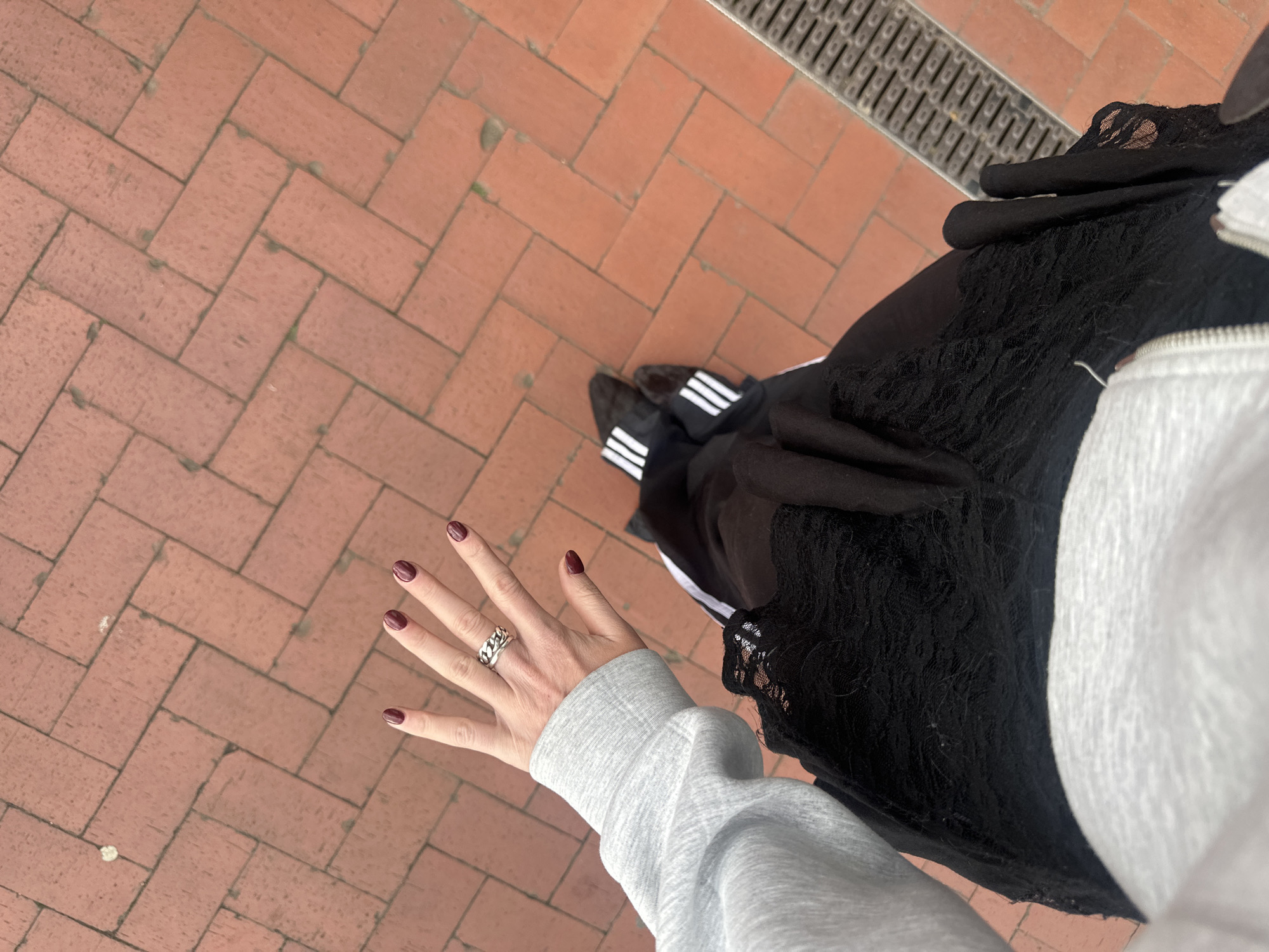 A street-level perspective shows a person standing on a herringbone-patterned brick pavement, typical of urban areas. The person's left hand is visible, displaying manicured nails painted in a dark red color and a prominent silver ring on the ring finger. The individual is wearing a gray, long-sleeved sweatshirt and a black skirt with lace detailing. Adidas-branded shoes with the signature three white stripes are visible. The environment includes a metal drainage grate at the edge of the pavement. The location is suggested to be Tunbridge Wells, United Kingdom, based on the typical urban design elements, but this cannot be confirmed from the image alone. The focus is on the person and their attire, with the environment providing context.