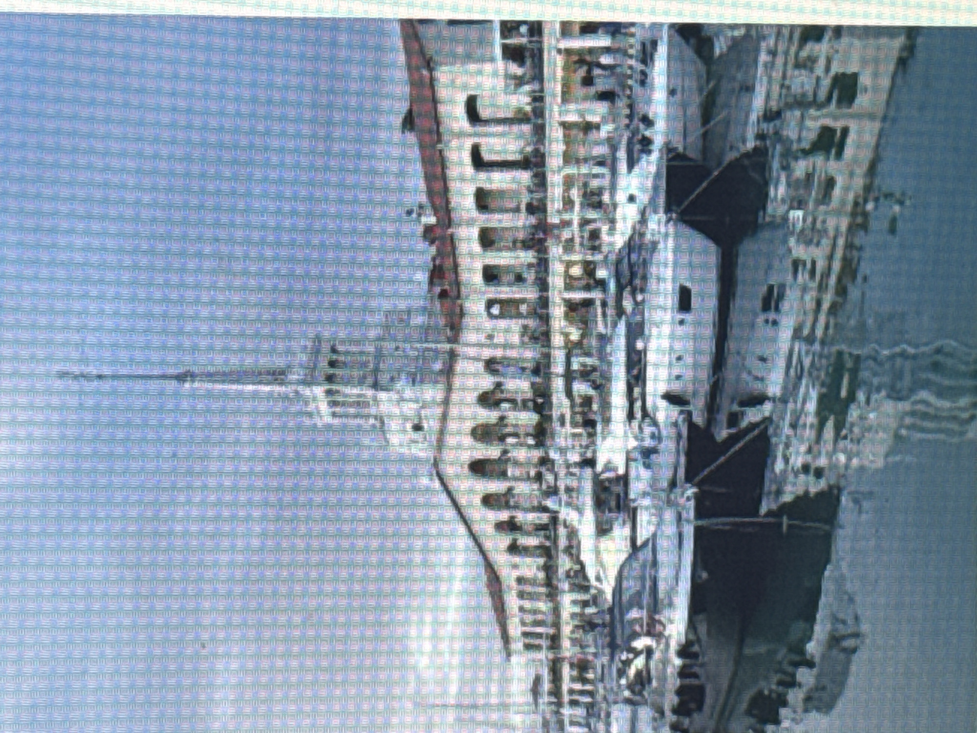 An exterior, eye-level, medium shot captures a marina with several yachts docked and a large building in the background under a clear sky. The building, a multi-story structure with a tall spire, features a white facade with arches and a reddish roof. The yachts vary in size and color, with some appearing dark and others white, and their reflections are visible on the calm water. The scene appears to be a sunny day, suggesting a leisurely or active environment at a port.