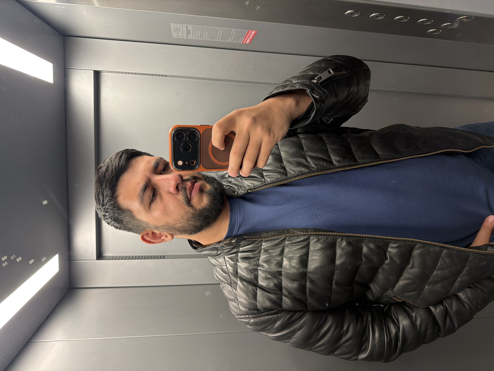 A man stands in an elevator, taking a selfie with his smartphone. He is wearing a blue shirt and a black quilted leather jacket. The elevator has a gray interior with overhead lighting. There are safety and informational stickers on the walls. The location is in a city, likely Krasnoyarsk, Russia, due to the prompt instruction, though specific location indicators within the image are absent.