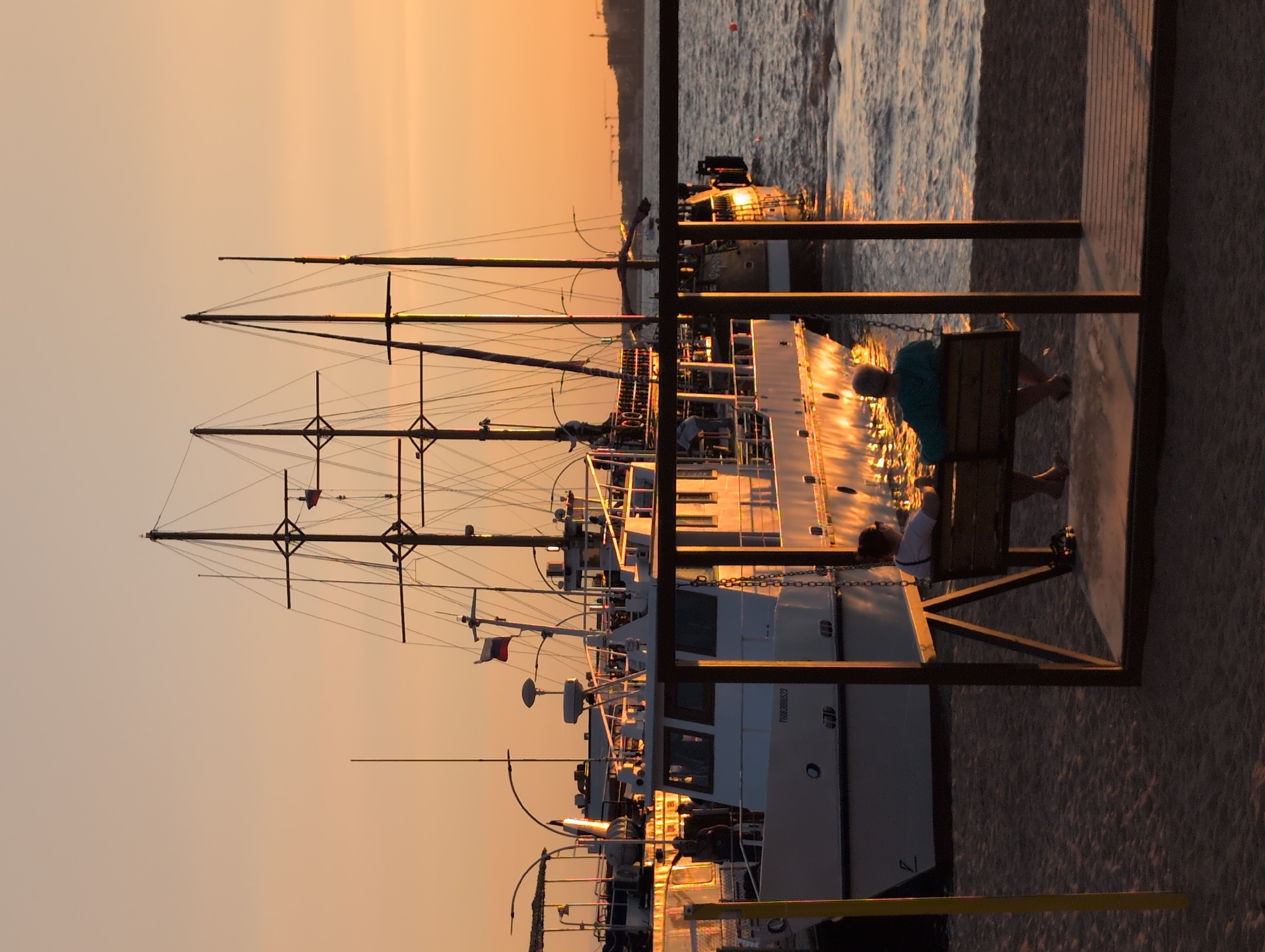 Two people are sitting on a swing on a sandy beach, facing the sea. The sun is setting, casting a warm golden glow over the scene. In the background, several boats are docked, including a large sailing ship with multiple masts and a smaller white vessel. The sea is calm with gentle ripples. The overall atmosphere is peaceful and serene, with the setting sun highlighting the boats and the water. This scene appears to be from the Gelendzhik embankment in Russia, known for its picturesque sunsets and harbor views.