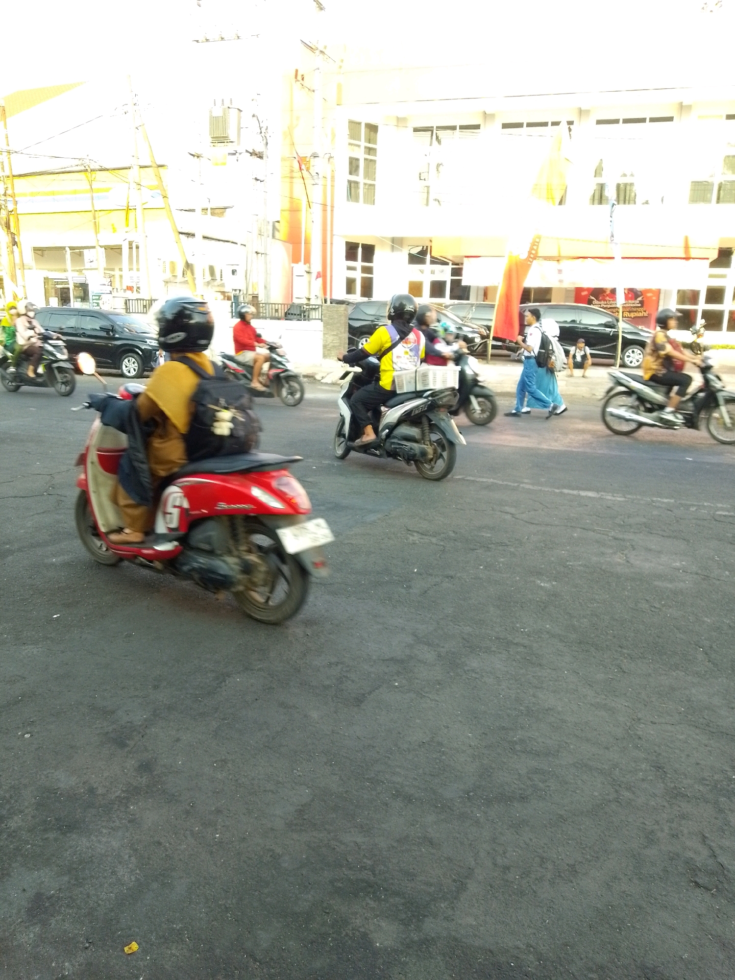 The image depicts a street scene in Jember, Indonesia, characterized by a clear sky and asphalt road. Several individuals on motorcycles are visible. In the foreground, a person wearing a yellow shirt and black helmet rides a red scooter. To the left, another person on a black motorcycle is partially visible. In the background, several buildings with windows line the street. A black car is parked on the left side of the road. Further in the background, a person in a blue uniform and another in a yellow shirt on a motorcycle are also present. The scene suggests a busy urban environment with common modes of transportation in Indonesia.