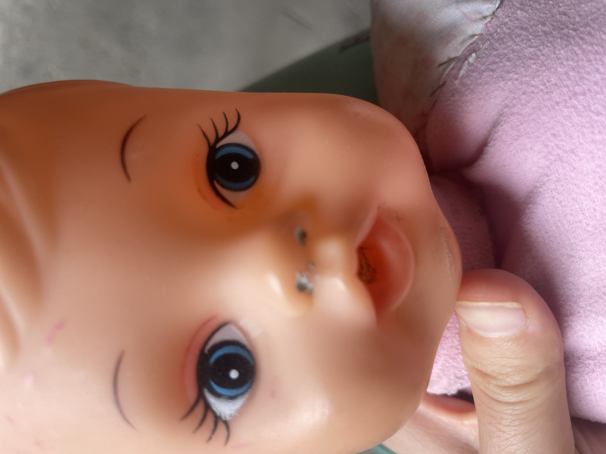 The image is a close-up of a doll's face, held by a human hand. The doll has a peach-colored plastic head with painted blue eyes, black eyelashes, and faint pink markings on its forehead. Its nose appears slightly smudged. The doll is wearing a pink garment, possibly a dress or outfit, with some stitching visible. A human finger is visible in the lower left portion of the image, supporting the doll's head. The background is out of focus and appears to be a neutral, indoor setting, possibly a wall or fabric. There is no information to indicate the location in Tashkent, Uzbekistan.