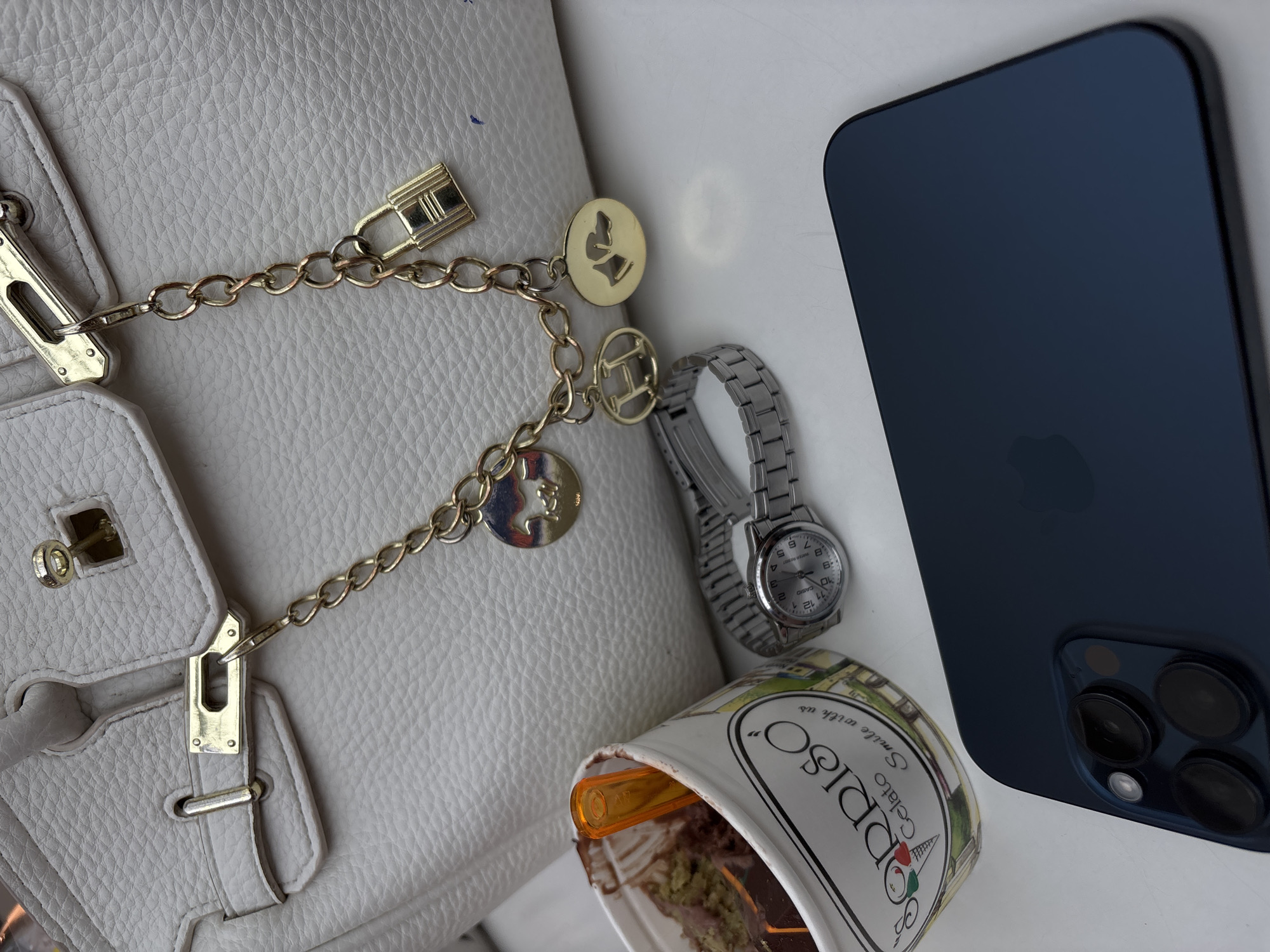 The image shows a still life on a white surface. A white leather handbag occupies the left side of the frame, with a gold chain and various charms attached. Next to the bag is a silver wristwatch. Nearby, there is a partially consumed container of "Sorriso Gelato." A dark blue smartphone with multiple camera lenses is positioned to the right. The location is unknown.