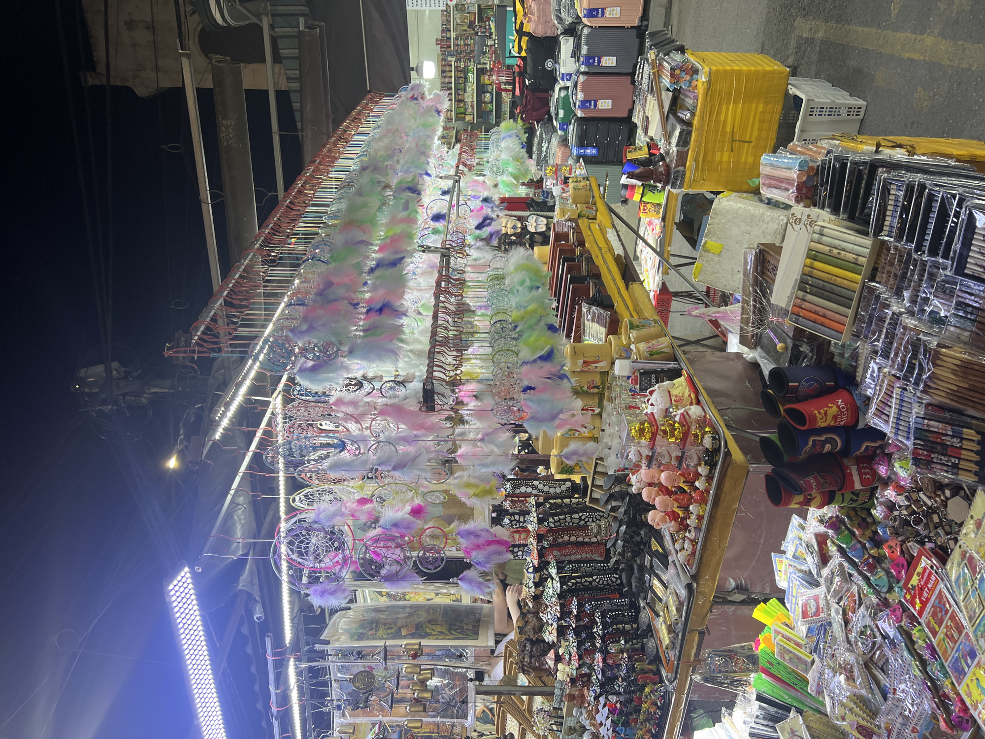 The image captures a vibrant night market scene in Hội An Ward, Vietnam. The primary subjects are a variety of goods displayed for sale, including numerous colorful dreamcatchers adorned with feathers, stacks of small suitcases, and an assortment of trinkets, souvenirs, and crafts. The setting is an outdoor market with overhead structures and bright artificial lighting illuminating the stalls. The activity is that of commerce, with vendors likely present, though no people are clearly visible or in focus. Notable details include the extensive display of dreamcatchers, suggesting they are a prominent item for sale. The lighting indicates it is nighttime. The overall impression is one of a lively and bustling market environment. There are some visible text elements, primarily on packaging and labels of the merchandise, though they are not legible enough to be transcribed.
