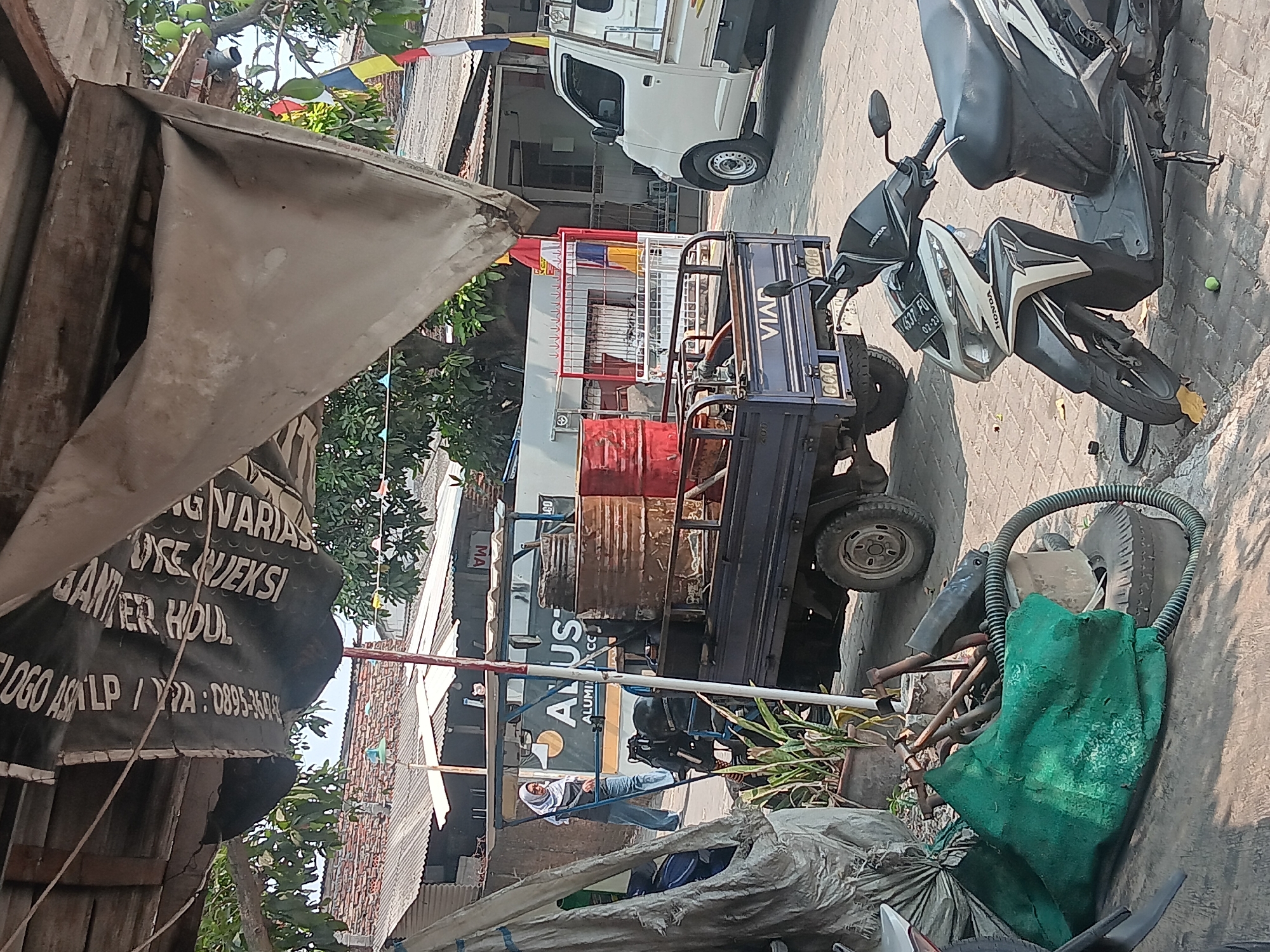 In Batursari, Indonesia, a street scene unfolds under a canopy of rough wooden beams supporting a weathered tarp. A black banner with white text hangs prominently, displaying what appears to be an advertisement with Indonesian writing, including "VARJASI" and "UDUL". To the left, a woman wearing a white hijab and blue jeans walks, carrying a dark bag. In the middle ground, a small blue truck with a metal frame, possibly a three-wheeled vehicle, is parked. It carries two large, rusty metal barrels and one red barrel. The truck has "VIAR" branding on its rear. To the right, a white pickup truck is parked, facing away from the viewer. In the foreground, a black and white scooter is parked on a brick-paved surface. Several green tarp-covered objects and some metal parts are scattered near the scooter. Buildings with corrugated metal roofs are visible in the background.