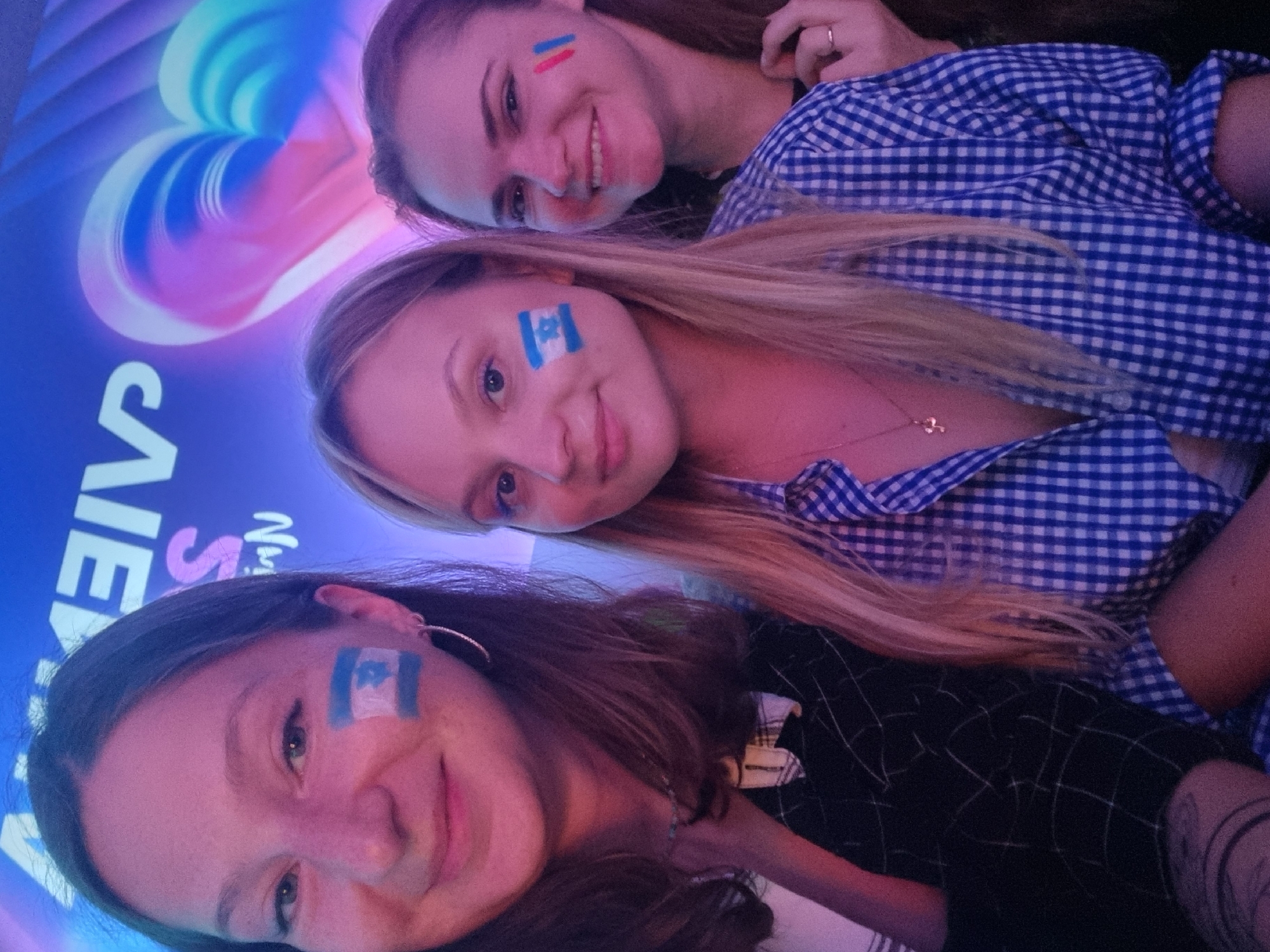 Three young women are posing for a selfie in Bat Yam, Israel. All three women have painted flags on their faces; two have Israeli flags and one has the French flag. The women are smiling. The background is blurred and features colorful, swirly designs and the word "CAINE". The image has a pink hue.
