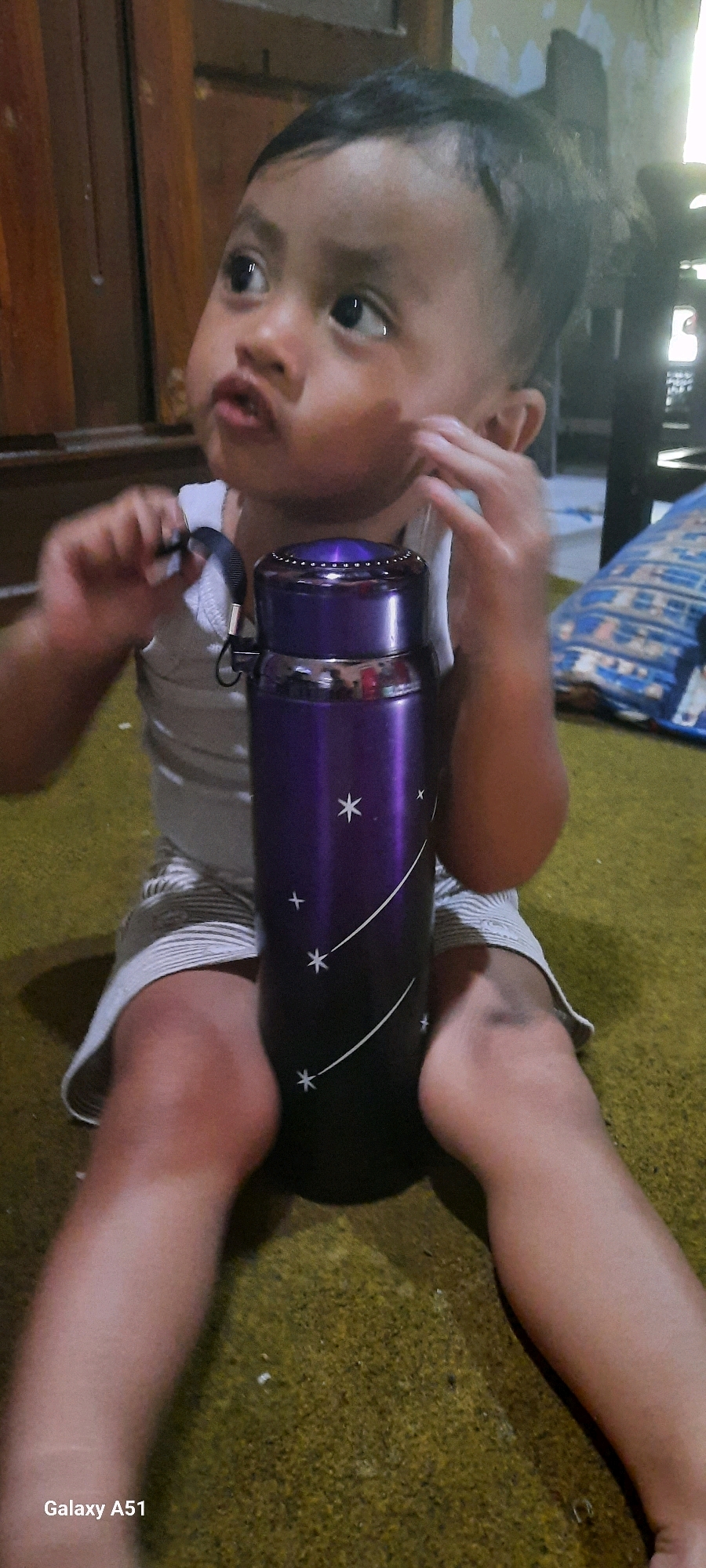 In an indoor setting, a young child is sitting on a textured, carpeted floor. The child is wearing a white tank top and striped shorts. They are holding a purple thermos bottle with white star-like patterns. The child's right hand is near their face, and their left hand is holding a strap from the thermos. In the background, there appears to be wooden furniture and a portion of a patterned cushion. The scene suggests a casual, domestic environment.