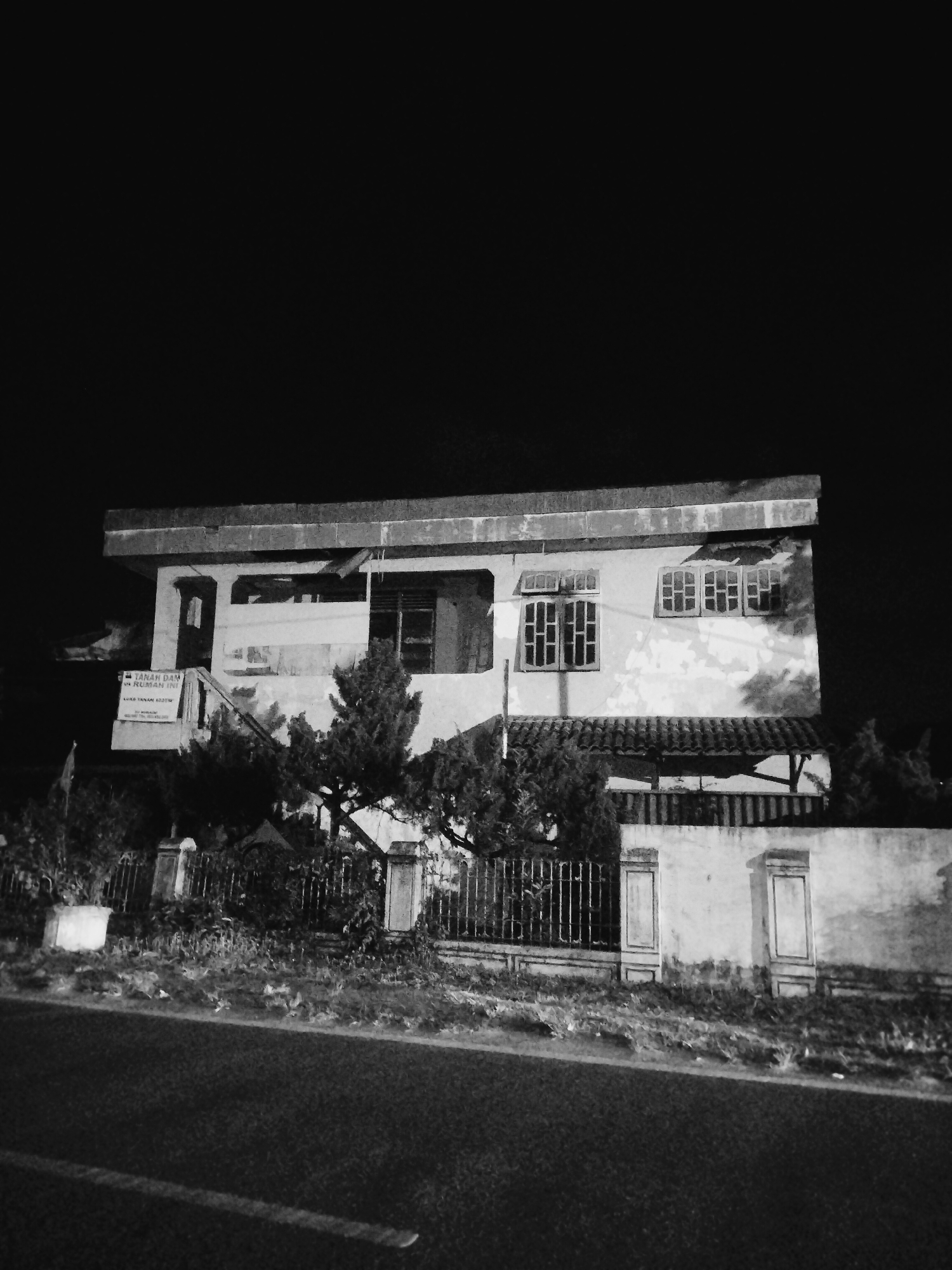 The photograph, taken in Lhokseumawe, Indonesia, presents a monochrome view of a two-story building at night. The sky is predominantly black, with the building illuminated by an unseen light source. The structure has a flat roof with visible deterioration on the exterior walls. Windows are visible on both floors, some with multiple panes. A sign is affixed to the building's facade. A fence and low wall border the property, with vegetation growing along the base of the wall. A road with a white line marker runs along the foreground.