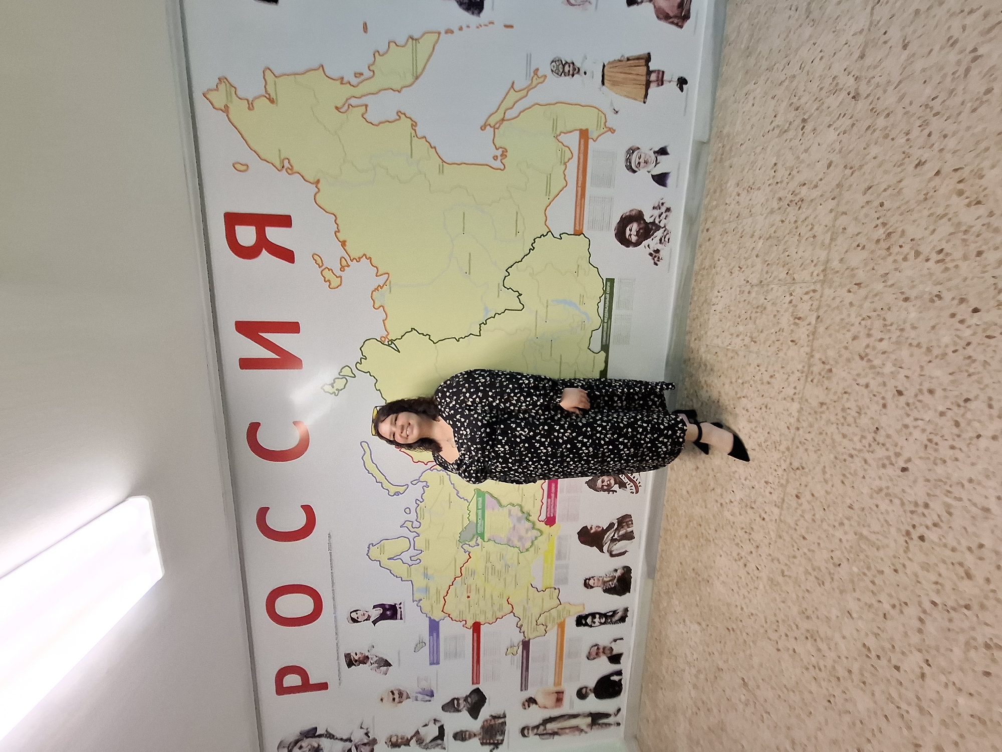 A woman is standing in front of a map of Russia. The map is posted on a wall next to a light fixture, with portraits on the right side. The floor is tiled. The word "РОССИЯ" is written in large red letters on the wall to the left of the map.