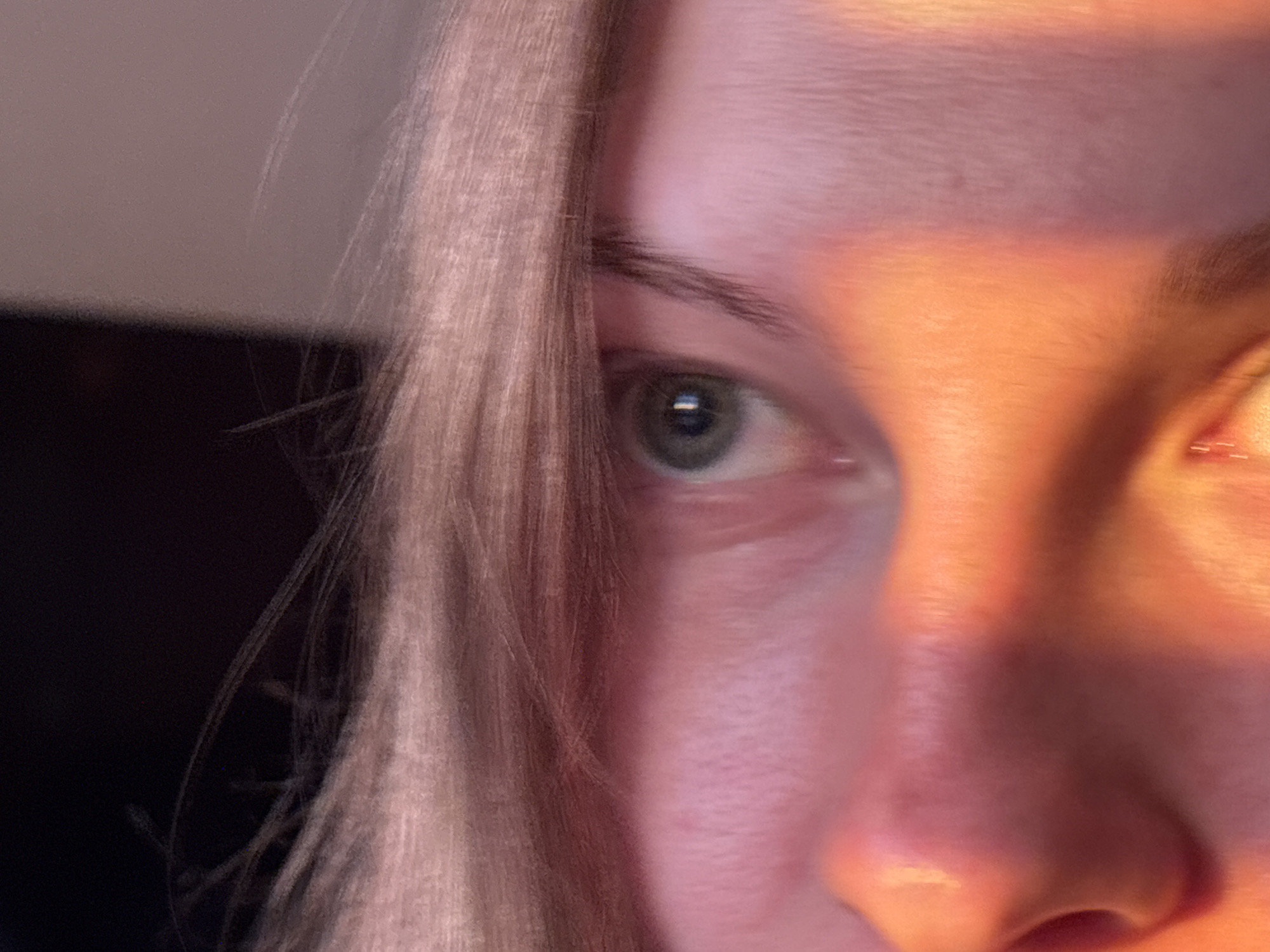The image presents a close-up, cropped forensic view of the upper left portion of a human face. The subject's left eye (viewer's left) is centrally focused and open, revealing a light-colored iris, possibly green or hazel, with a distinct black pupil. A rectangular reflection, indicative of a light source, is visible within the pupil. The eyebrow above the left eye is dark and well-defined. Strands of light-colored hair, appearing blonde or light brown, cascade down the left side of the face, partially obscuring the temple and cheek area.
The right side of the face (viewer's right), including the bridge and tip of the nose, is intensely illuminated by a strong, warm, orange-yellow light, creating a significant contrast with the cooler, shadowed tones on the left side. The skin exhibits texture, particularly around the eyes and on the forehead, ranging in color from pale pink to a saturated orange-red due to the direct illumination. The right eye is mostly obscured by the strong light and the composition.
The background on the far left is dark and out of focus, suggesting an indoor setting without identifiable objects or landmarks. There is a faint, lighter, out-of-focus area in the upper left background. The overall scene is dominated by the dramatic lighting, which highlights facial features and creates a pronounced chiaroscuro effect. There are no identifiable actions or interactions depicted. The image does not provide any visual information to determine a specific geographical location, including the city of Perm, Russia.