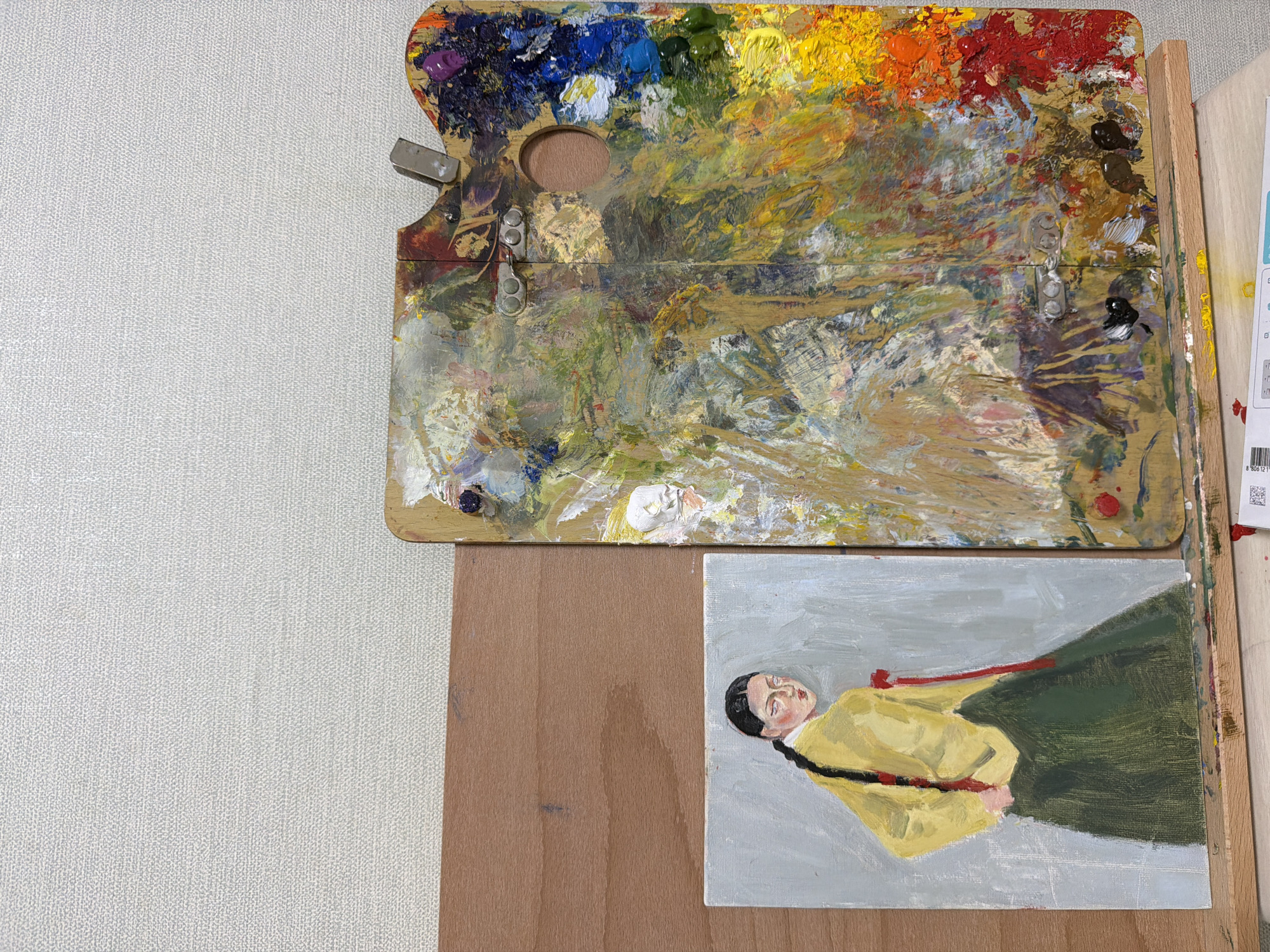 The image shows an artist's workspace. On the left is a small painting of a woman in traditional Korean clothing, a yellow top with a red sash and a dark green skirt. She has her back to the viewer, looking over her shoulder with a neutral expression. Her dark hair is braided. The painting is on a light gray background.
To the right of the painting is a large wooden artist's palette, heavily laden with a vibrant and thick mixture of oil paints in various colors including red, orange, yellow, blue, green, white, and brown. The palette itself is worn and appears to be hinged in the middle.
The workspace is set against a neutral, textured background, possibly a wall or canvas. The scene is lit by overhead light, casting subtle shadows. There is no indication of the city of Dongducheon-si, South Korea.