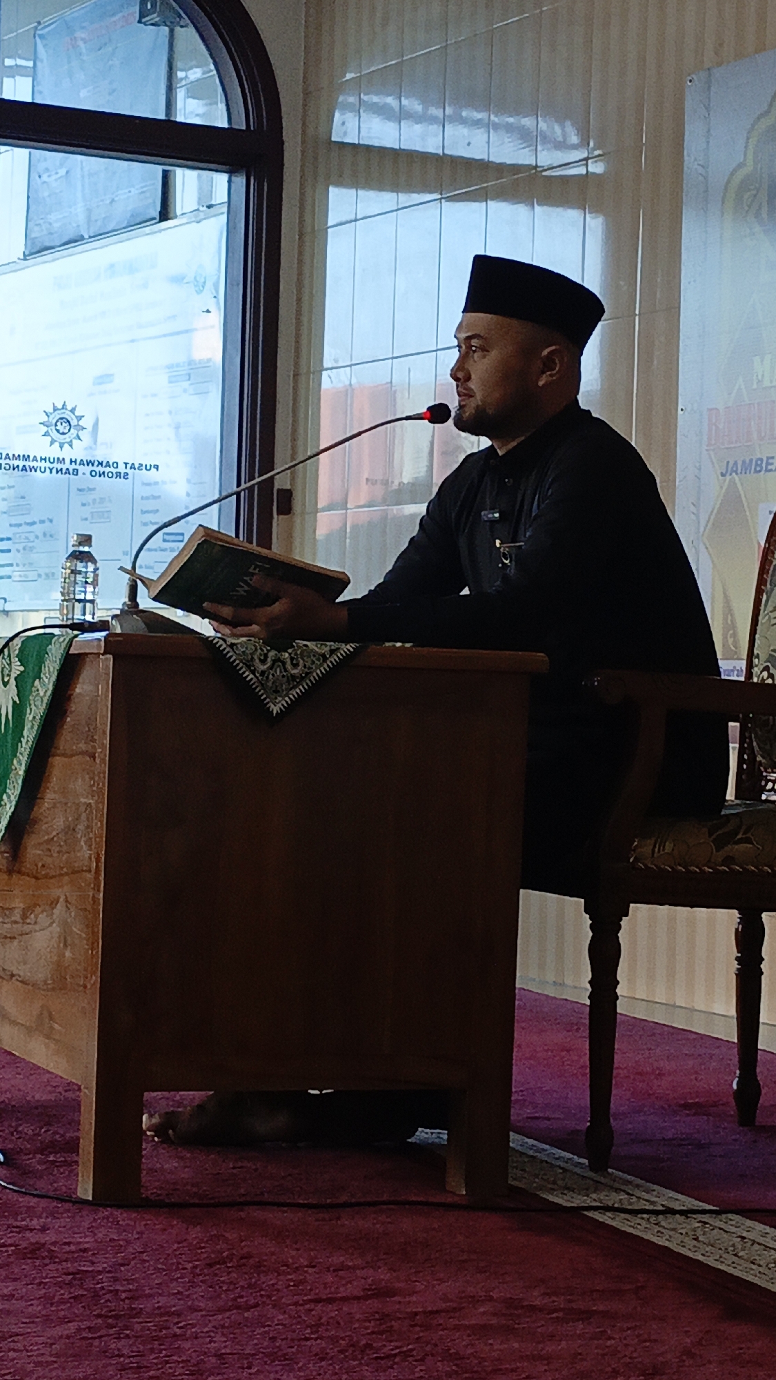 An Indonesian man wearing a black peci hat and a black long-sleeved shirt is seated at a wooden podium, reading from a book. He is facing the right side of the frame and is positioned behind a microphone. To his left on the podium is a water bottle. The background shows a large window with reflections and a banner with text. The man's legs are not visible below the podium, but his feet are visible underneath the podium, one resting on the maroon carpet. The scene appears to be an indoor event, possibly a lecture or religious service, located in Srono, Indonesia, given the context of the image.