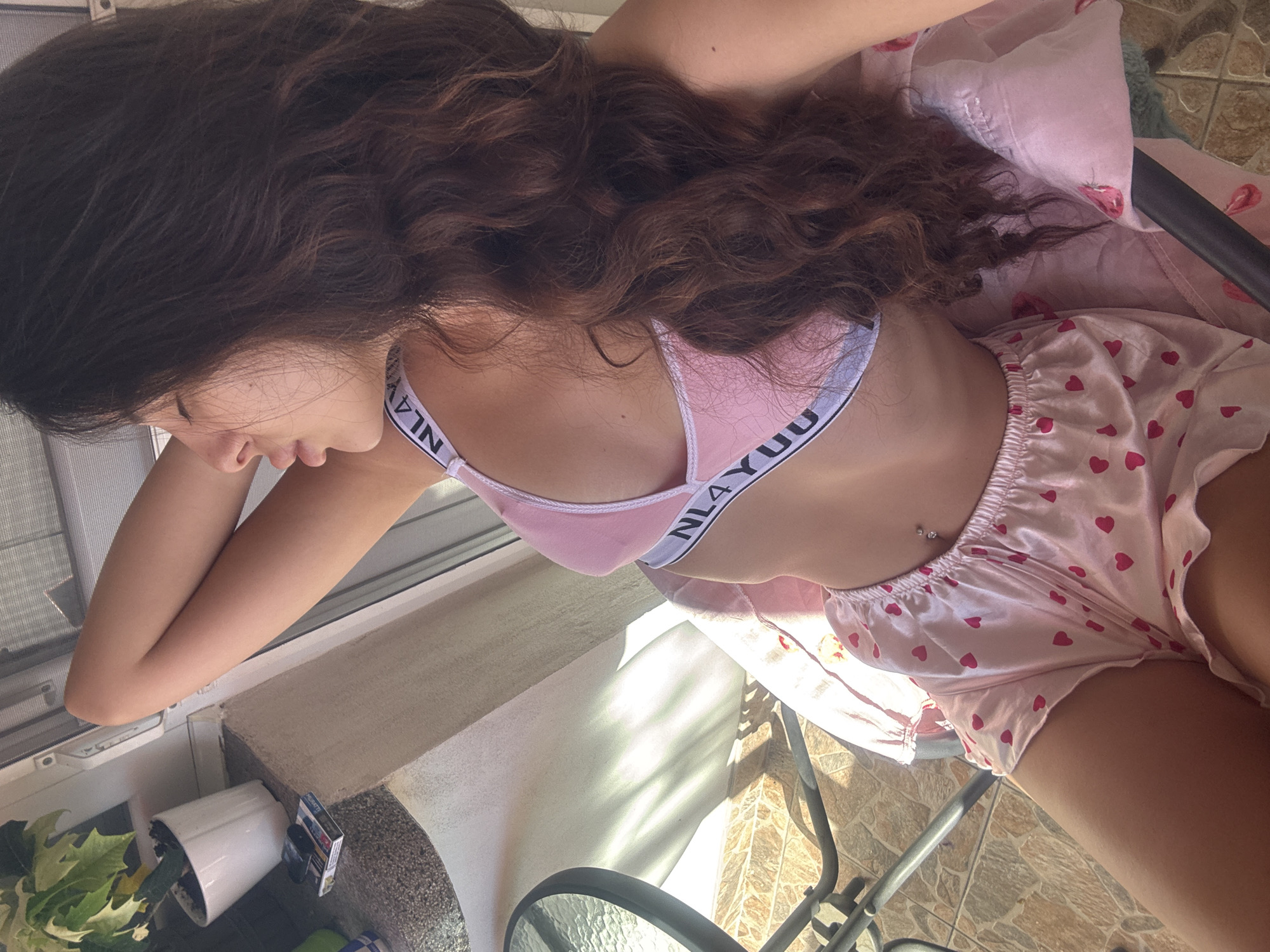 A young woman with long, dark, wavy hair is posing for a selfie on an outdoor patio. She is wearing a pink bralette with black lettering that says "NL4YUU" and pink satin shorts with a heart pattern. She has a piercing on her navel. The background shows a table and chair, a plant in a white pot, and a tiled wall. The lighting suggests it is daytime, possibly morning or afternoon, with sunlight casting shadows. The scene is likely in a residential area, given the patio setting.