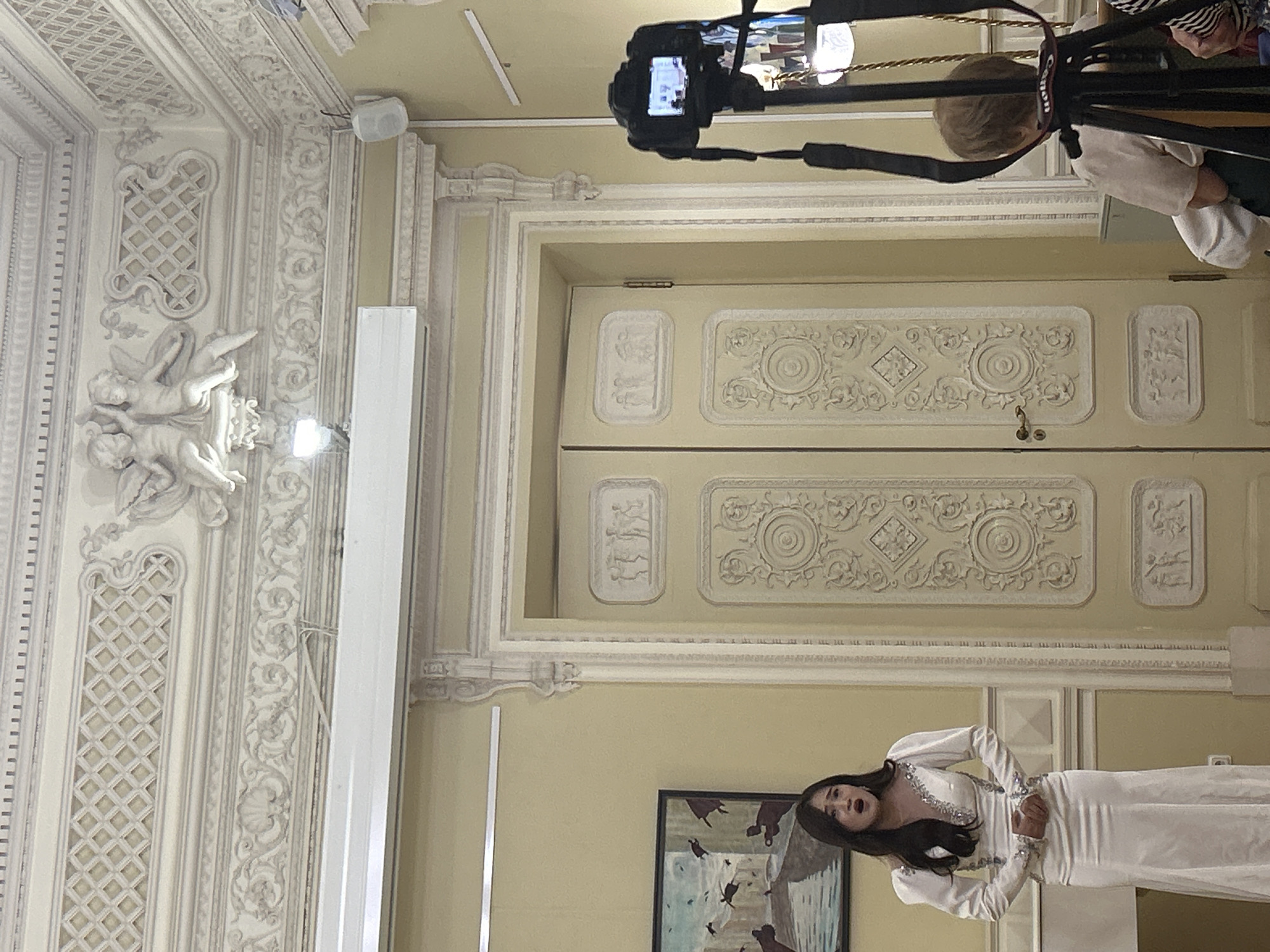 In a room with ornate white molding and a pale yellow wall, a woman in a white gown stands on the left, her mouth open as if singing. She has long dark hair and is wearing a v-neck dress with beaded detailing on the bodice and sleeves. To her right, a large, light-colored double door is set into the wall, featuring decorative paneling and relief carvings. Above the door, elaborate molding with cherubic figures adorns the ceiling. A black DSLR camera on a tripod is positioned in the foreground on the right, facing the woman. Part of a person's head and shoulders, viewed from the back, is visible behind the tripod. A framed painting depicting birds in flight hangs on the wall to the left of the woman. The overall impression is of a performance or recording taking place in a formal, decorated interior space.