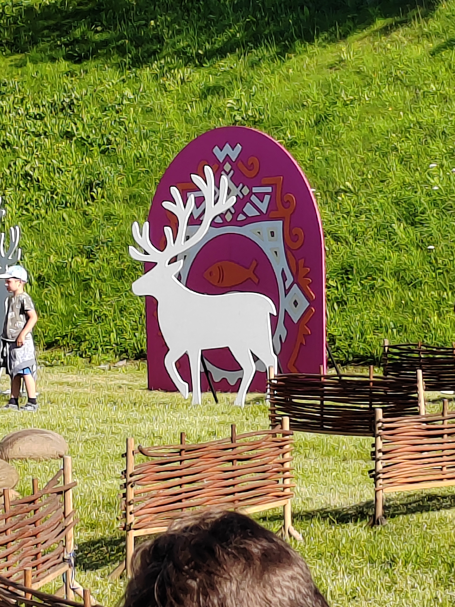 In an outdoor grassy area, a white cutout of a deer with large antlers stands in front of a purple, arched decorative panel. The panel features geometric patterns in light blue and orange, with a stylized orange fish. To the left of the deer, a young boy wearing a gray t-shirt, shorts, and a white baseball cap stands facing the deer cutout. In the foreground, there are sections of rustic, woven wooden fences. The background consists of lush green grass, with some dappled sunlight creating shadows. The scene appears to be set up as a decorative display or part of an event in an unspecified urban or suburban park.